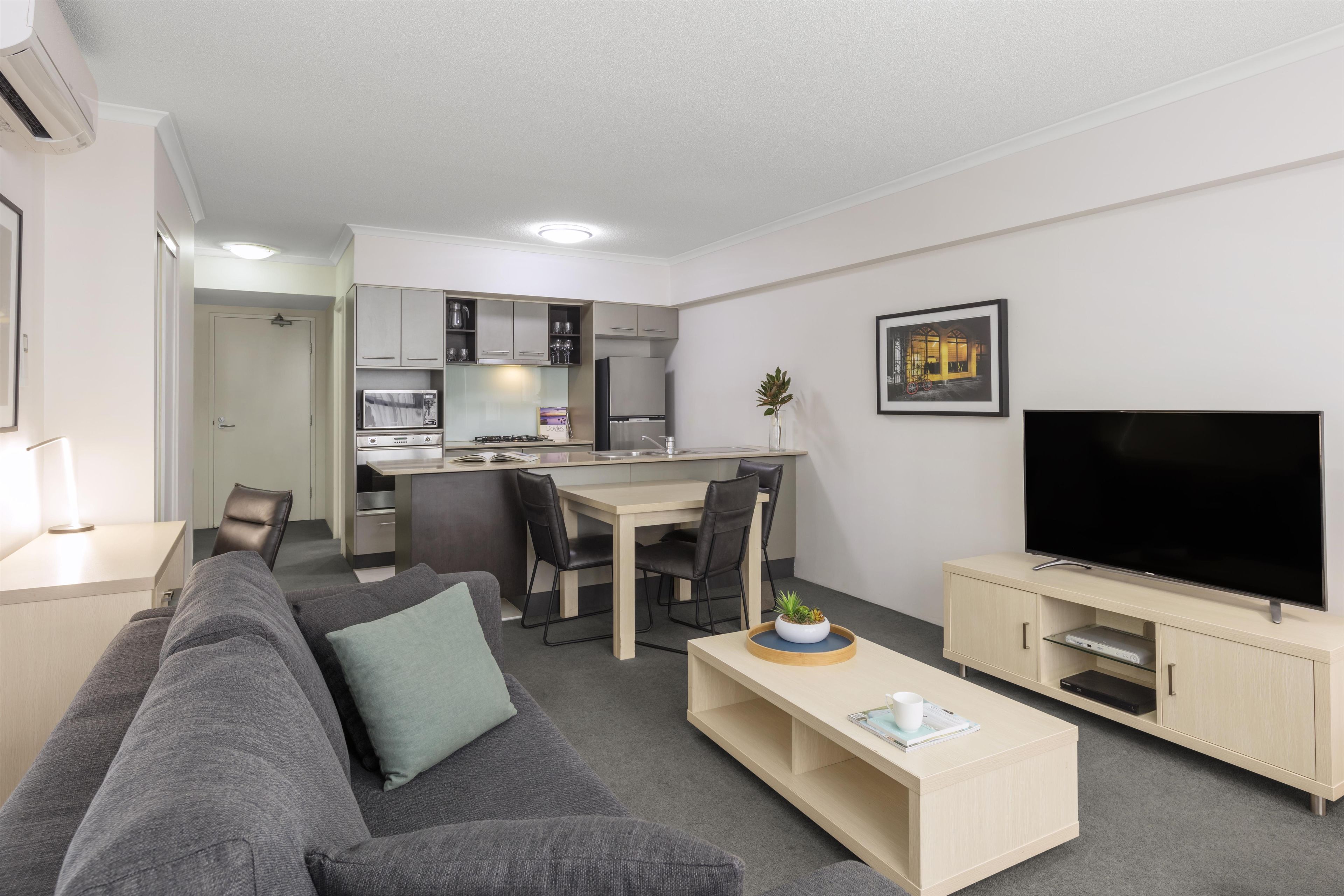 A modern living room with a grey sofa, a coffee table, a TV, and a kitchen area with cabinets and appliances.