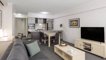 A modern living room with a grey sofa, a coffee table, a TV, and a kitchen area with cabinets and appliances.
