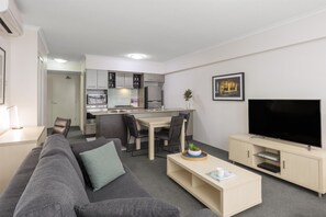 A modern living room with a grey sofa, a coffee table, a TV, and a kitchen area with cabinets and appliances.