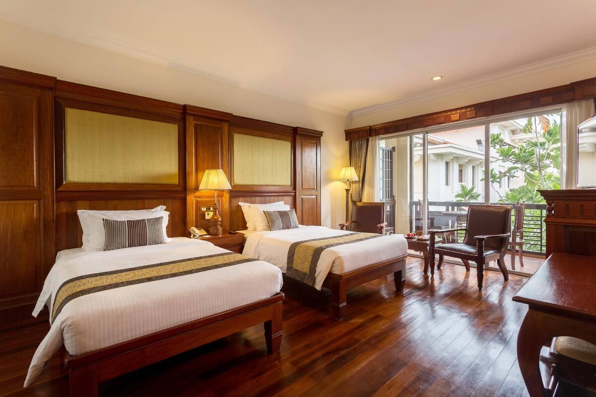 Habitación de hotel con dos camas de madera, un escritorio de madera, una silla y un balcón con vistas a la vegetación.