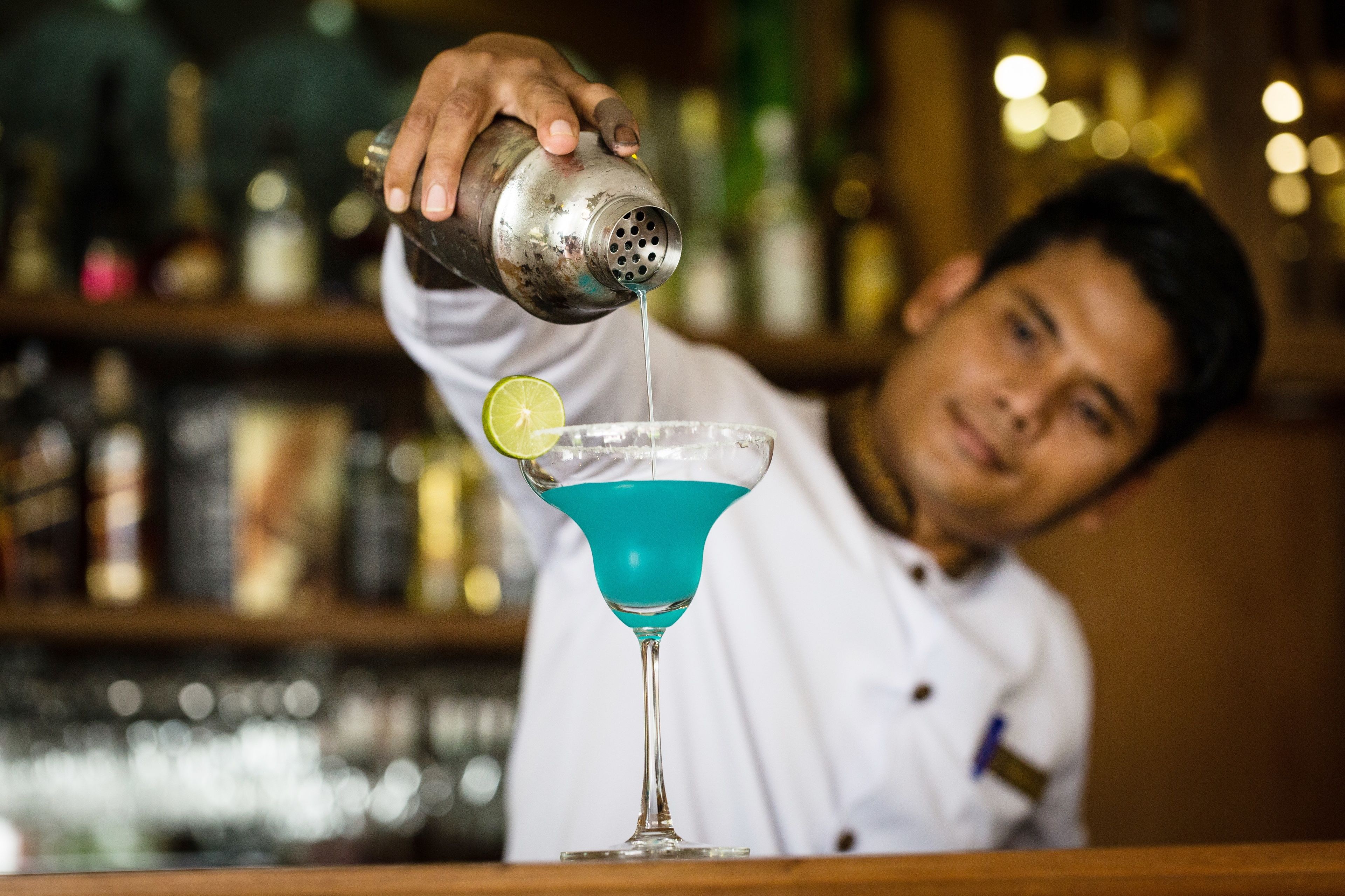 A bartender pouring a blue cocktail into a martini glass with a lime wedge.
