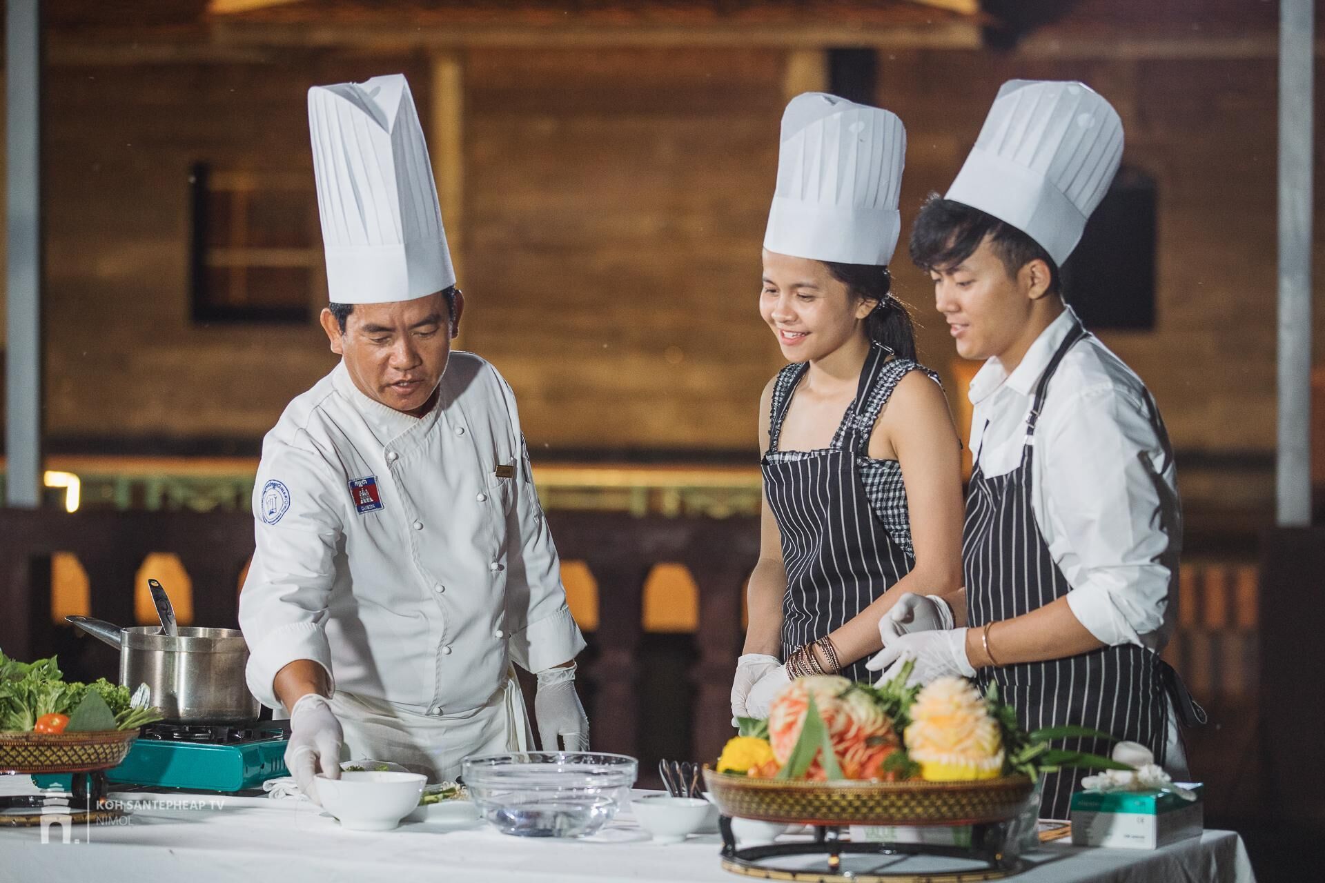 Three chefs in a kitchen setting with a table set for a meal, including bowls, plates, and a pot.