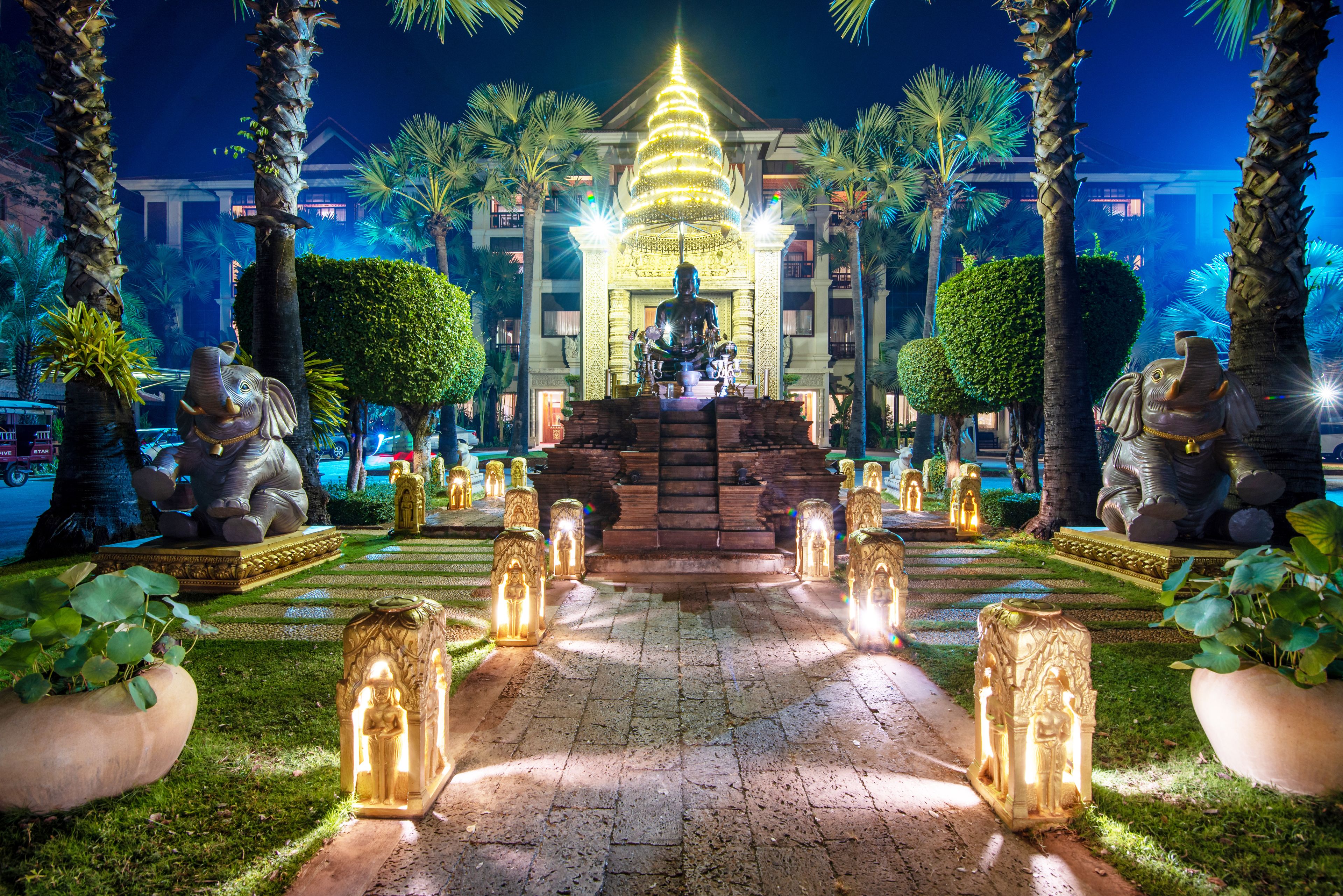 A nighttime view of a temple-like structure with a central statue, surrounded by palm trees and illuminated by lights.