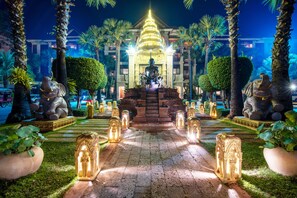 Una vista nocturna de una estructura tipo templo con una estatua central, rodeada de palmeras e iluminada por luces.