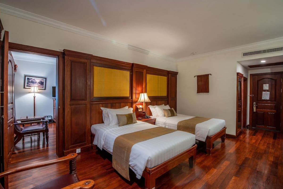 Habitación de hotel con dos camas, suelo de madera, un escritorio con silla, una lámpara y un cuadro en la pared.
