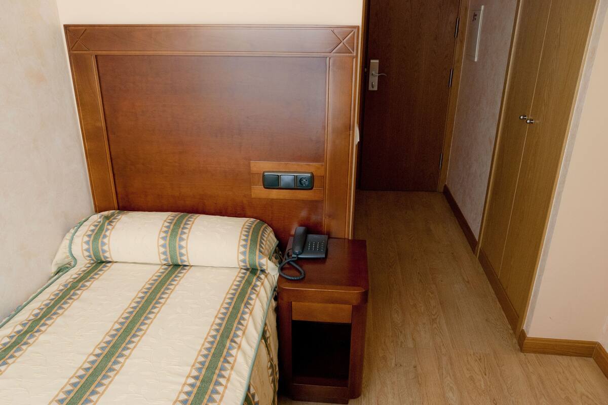 Una cama bien tendida con una colcha estampada, una mesita de noche con teléfono y una puerta de madera al fondo.