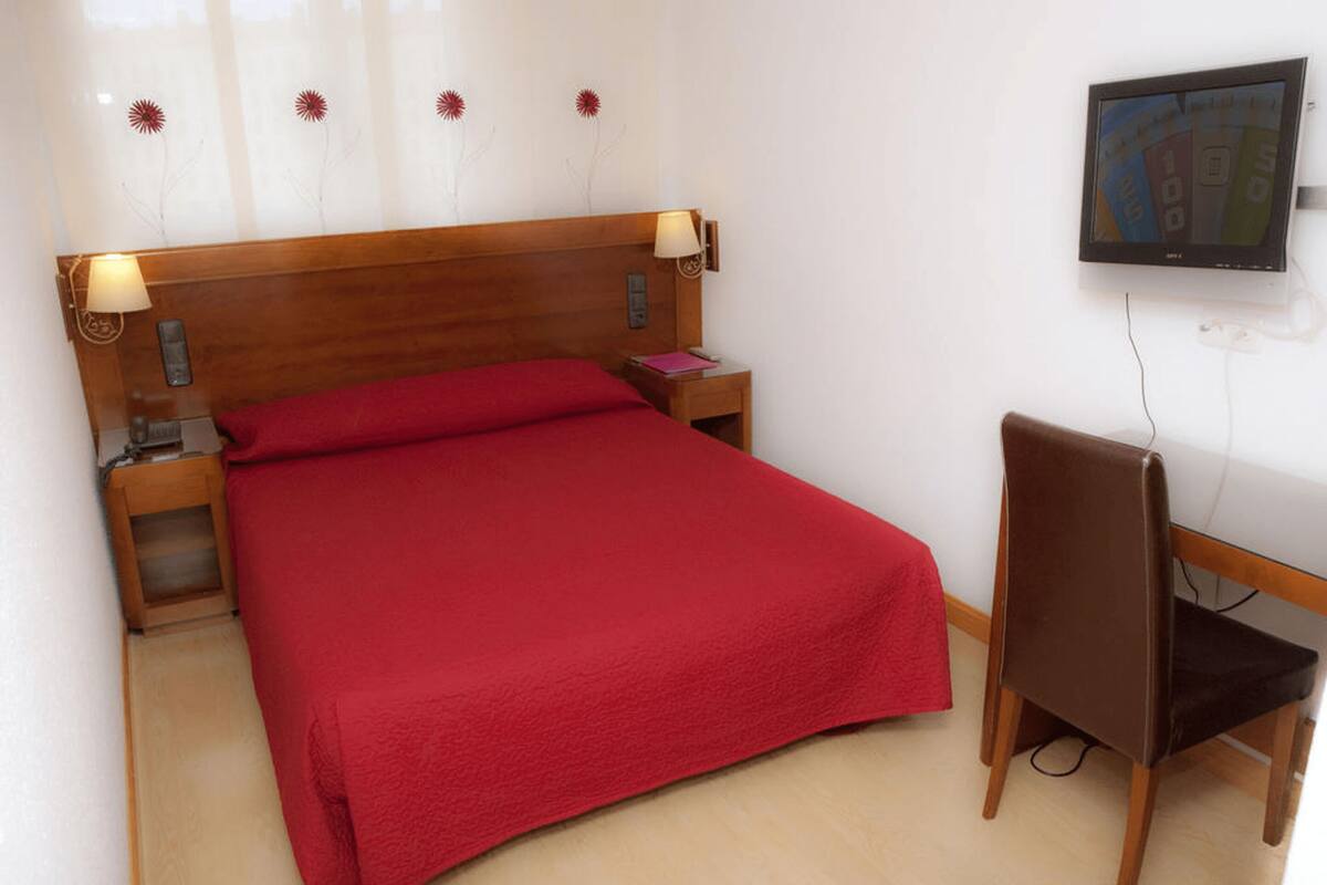 Una cama bien hecha con una manta roja, un cabecero de madera, lámparas de noche, una mesita de noche pequeña, una silla y un televisor montado en la pared.