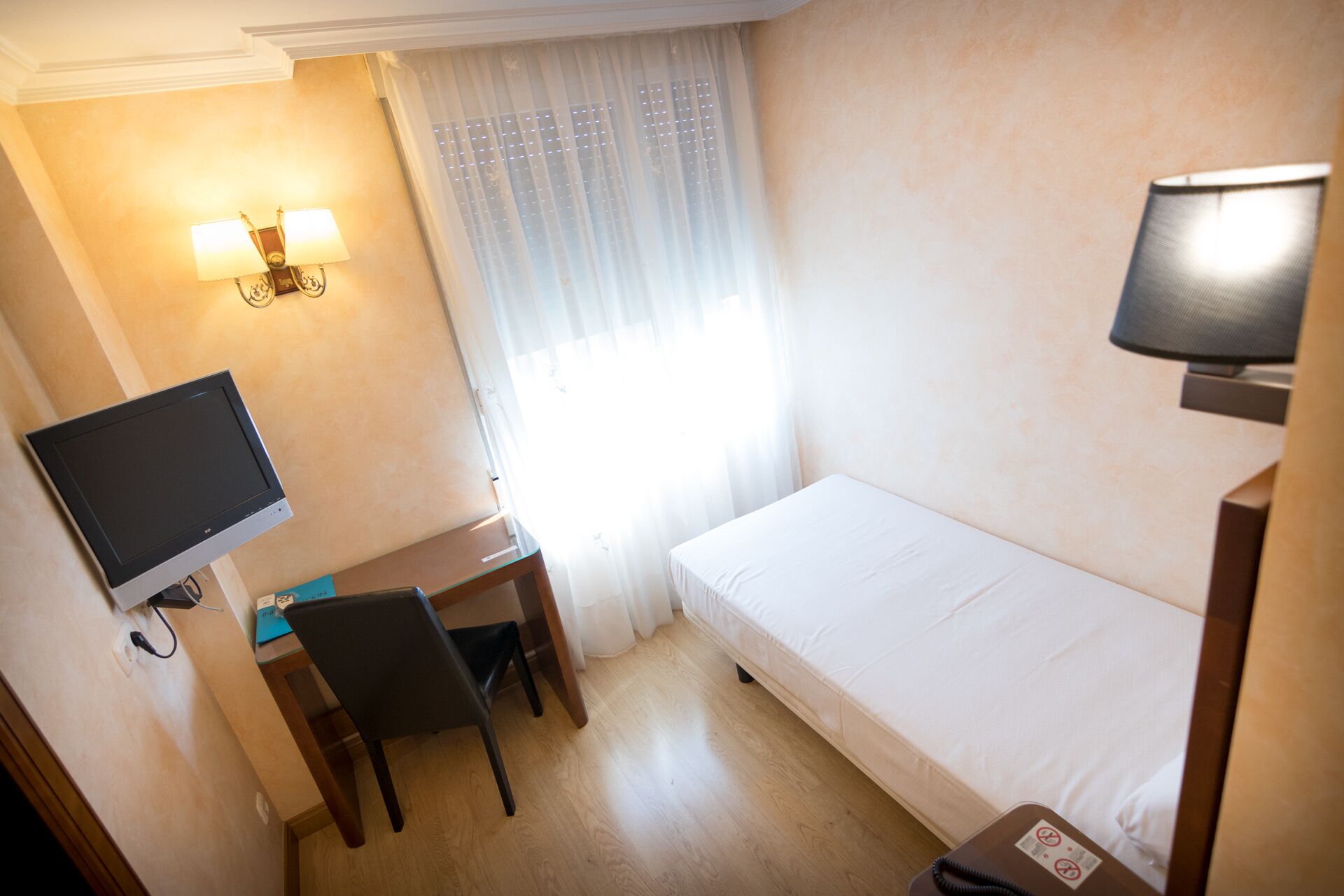 Una camera d'albergo piccola con un letto singolo, una scrivania con una sedia, una televisione e una finestra con tende leggere.