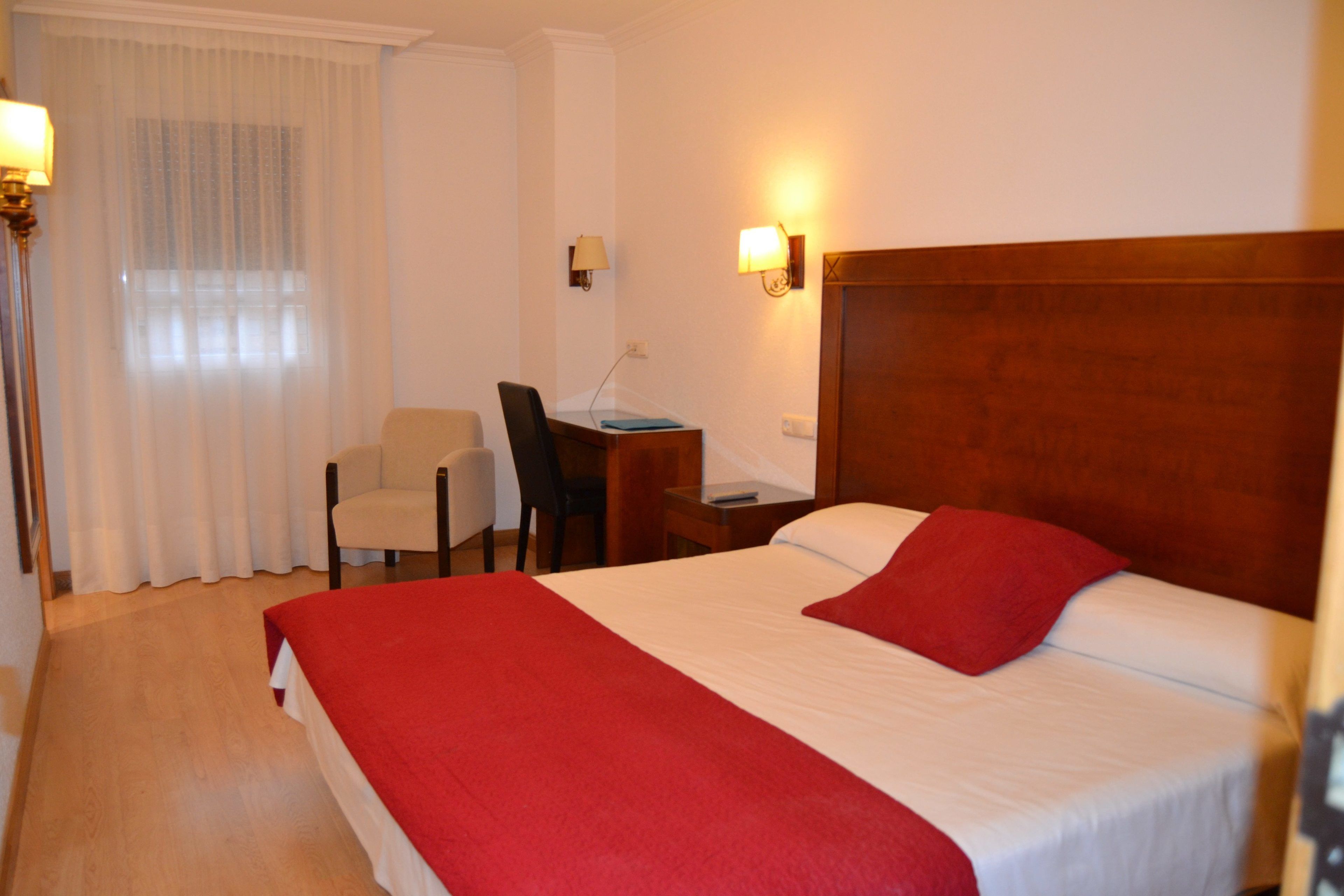 A hotel room with a large bed, a desk with a chair, a lamp, and a window with curtains.