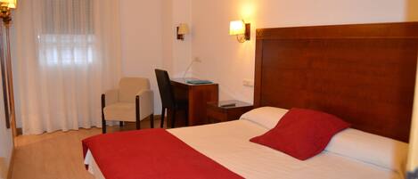 Habitación de hotel con una cama grande, un escritorio con una silla, una lámpara y una ventana con cortinas.