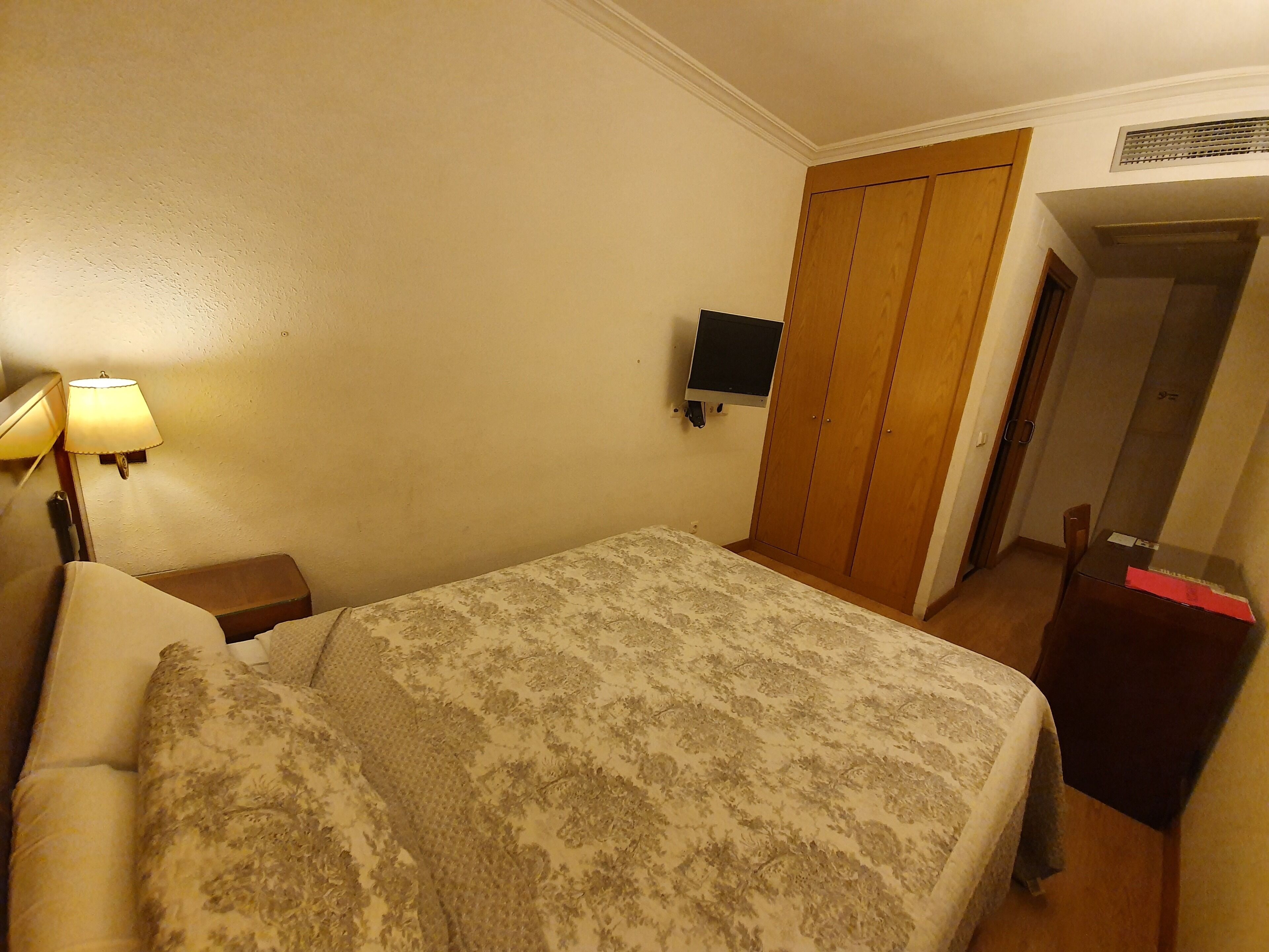 Un dormitorio con cama, mesita de noche con lámpara, televisor montado en la pared y armario.