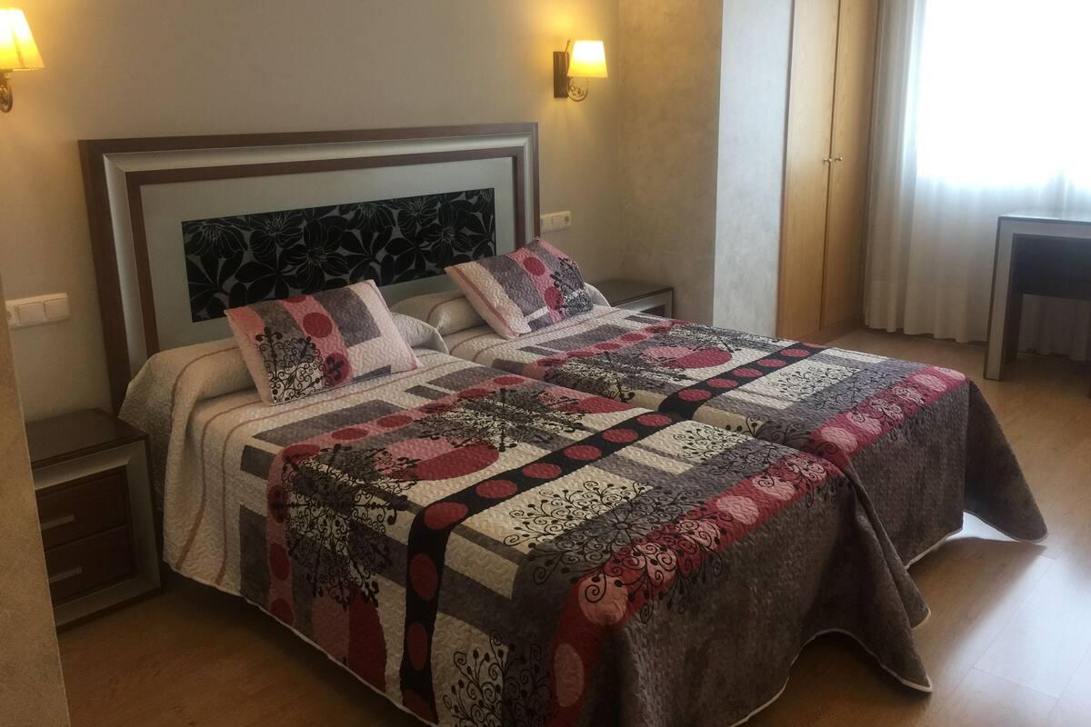 Una cama bien hecha con colcha estampada, mesitas de noche con lámparas y vista a una ventana con cortinas.