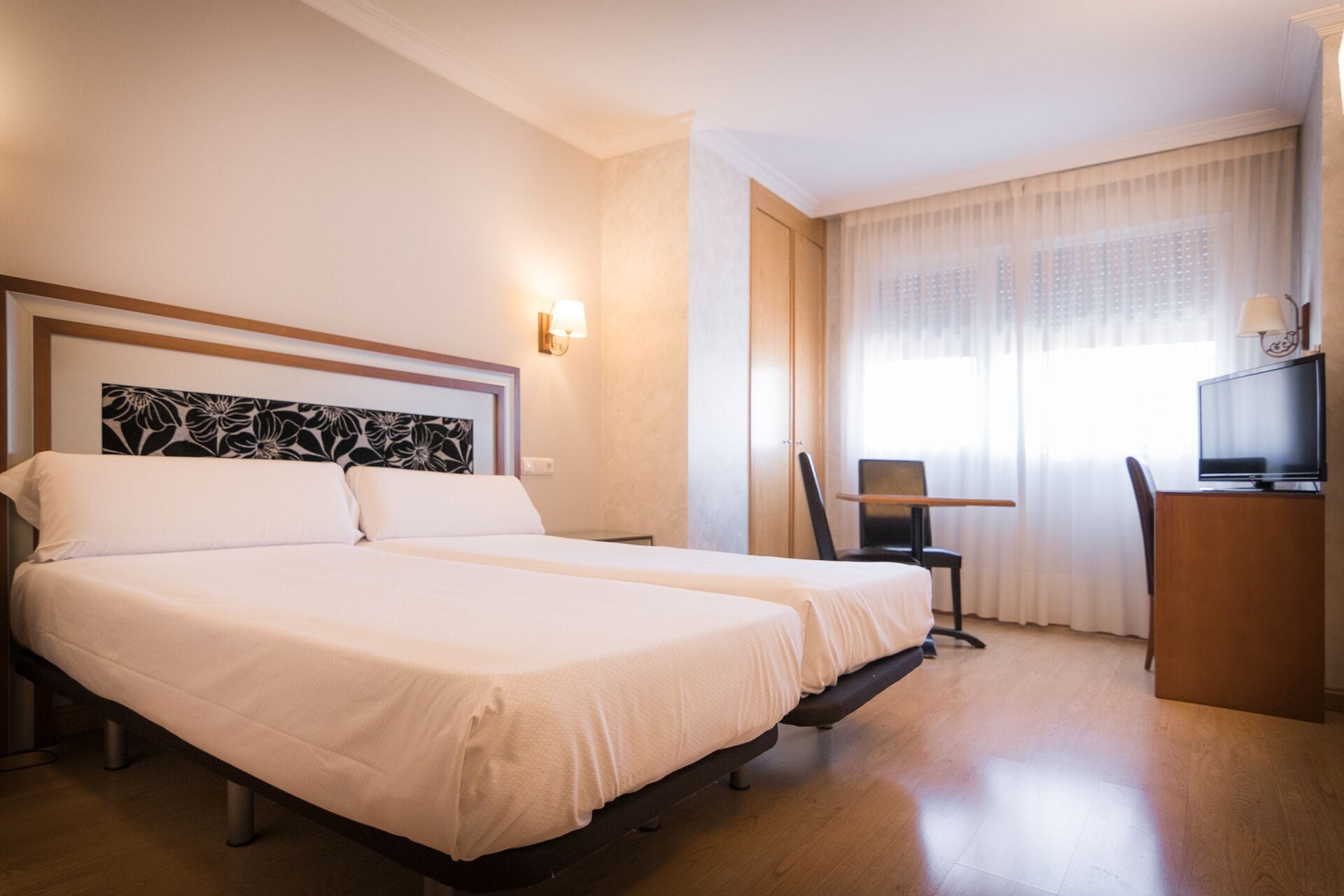A hotel room with two beds, a desk, a chair, a television, and a window with curtains.