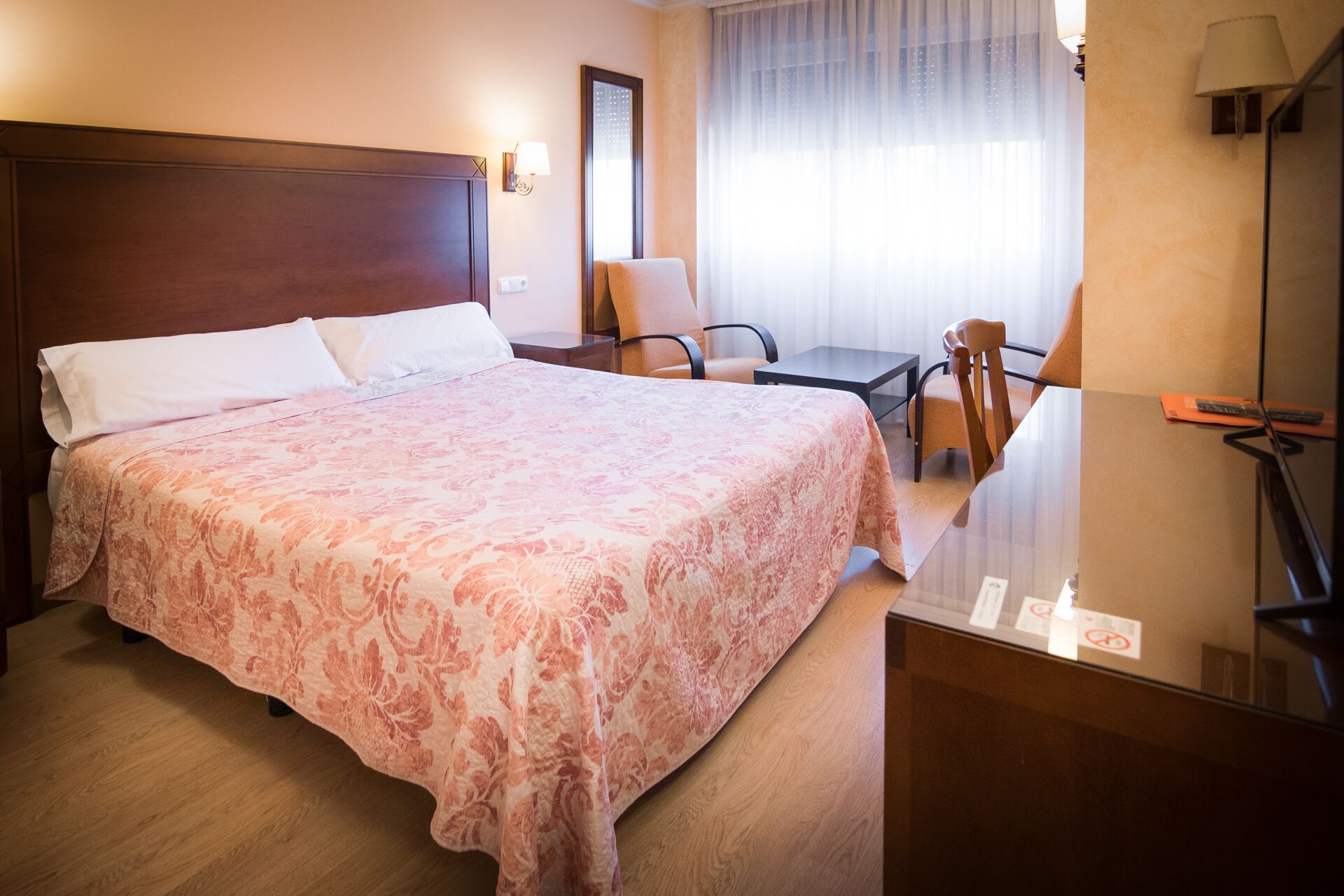 Habitación de hotel con una cama grande, un escritorio, dos sillas, una lámpara y una ventana con cortinas.