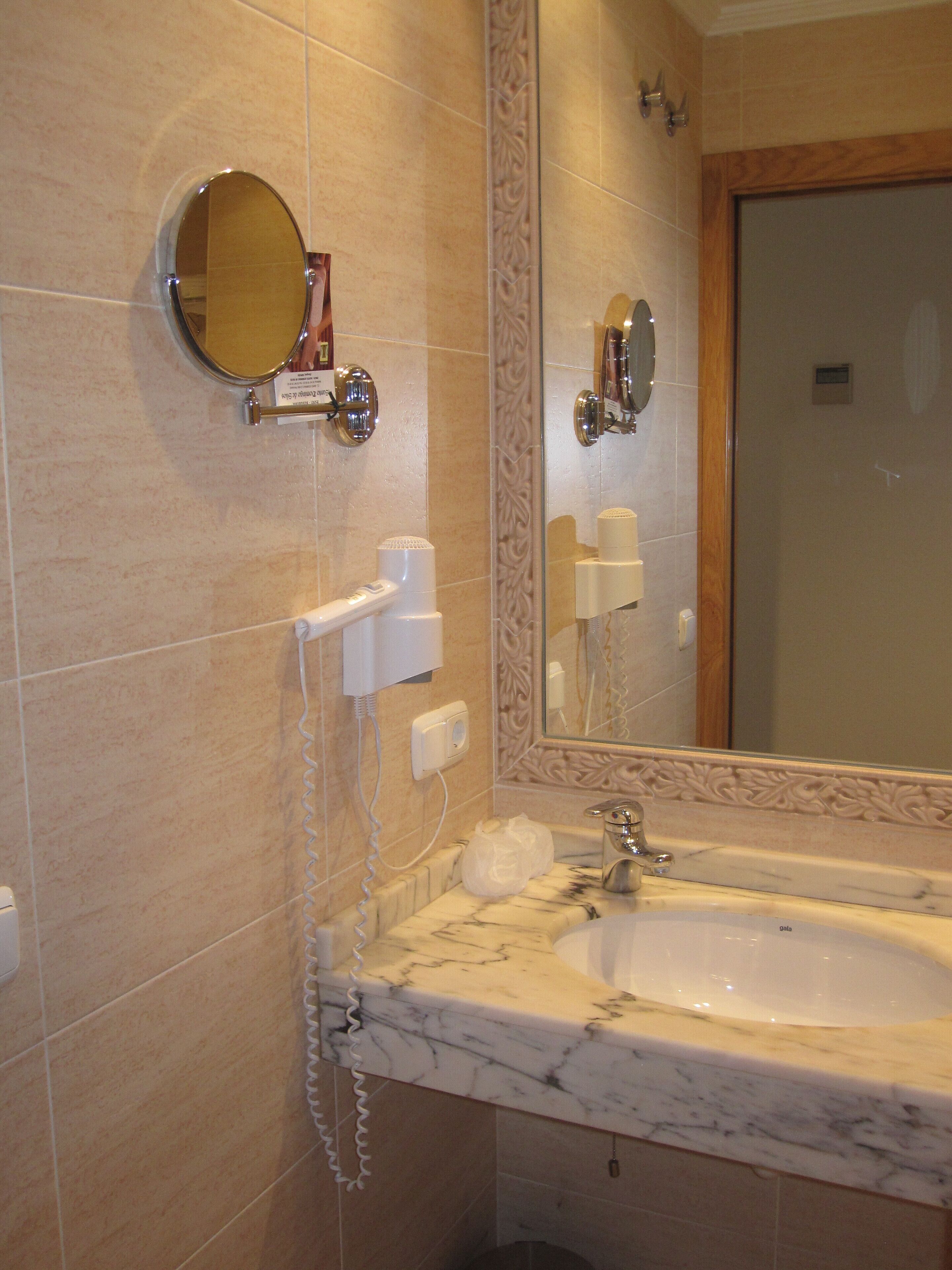 A bathroom with a marble sink, a mirror, a round mirror, a hairdryer, and a towel dispenser.