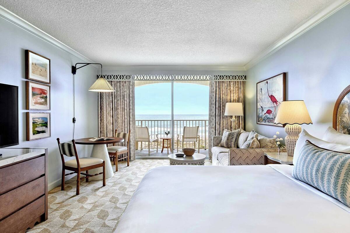 A hotel room with a large bed, a desk with a chair, a small table, a sofa, and a balcony with a view of the sea.