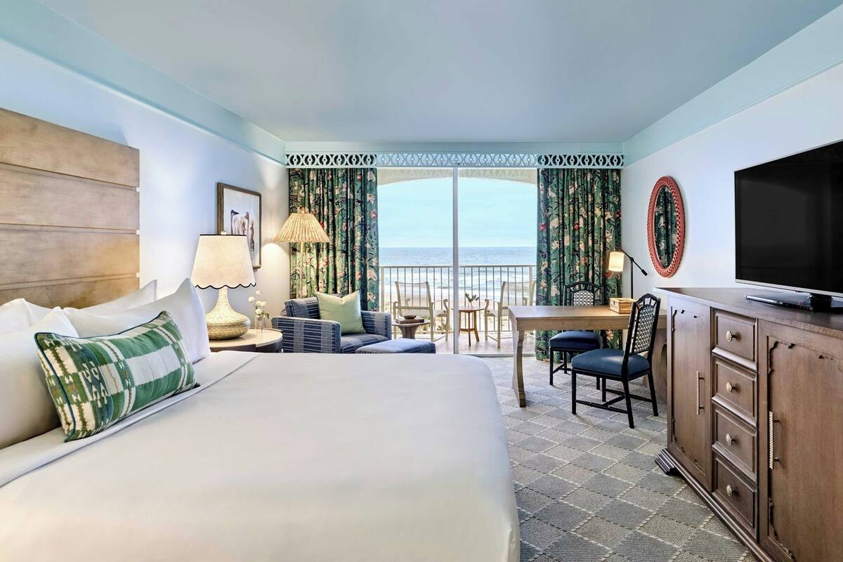 A hotel room with a large bed, a desk, a chair, a TV, and a view of the ocean.