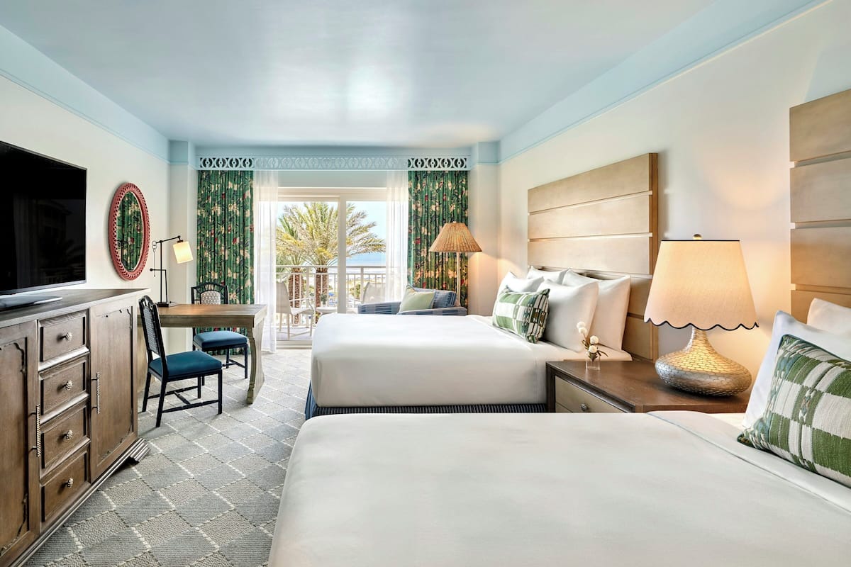 A hotel room with two beds, a desk, a TV, and a view of the outdoors.