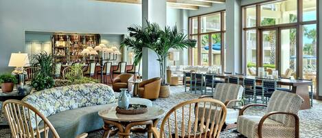 A spacious lobby with a high ceiling, large windows, a central seating area with sofas and chairs, and a bookshelf filled with books and decorative items.