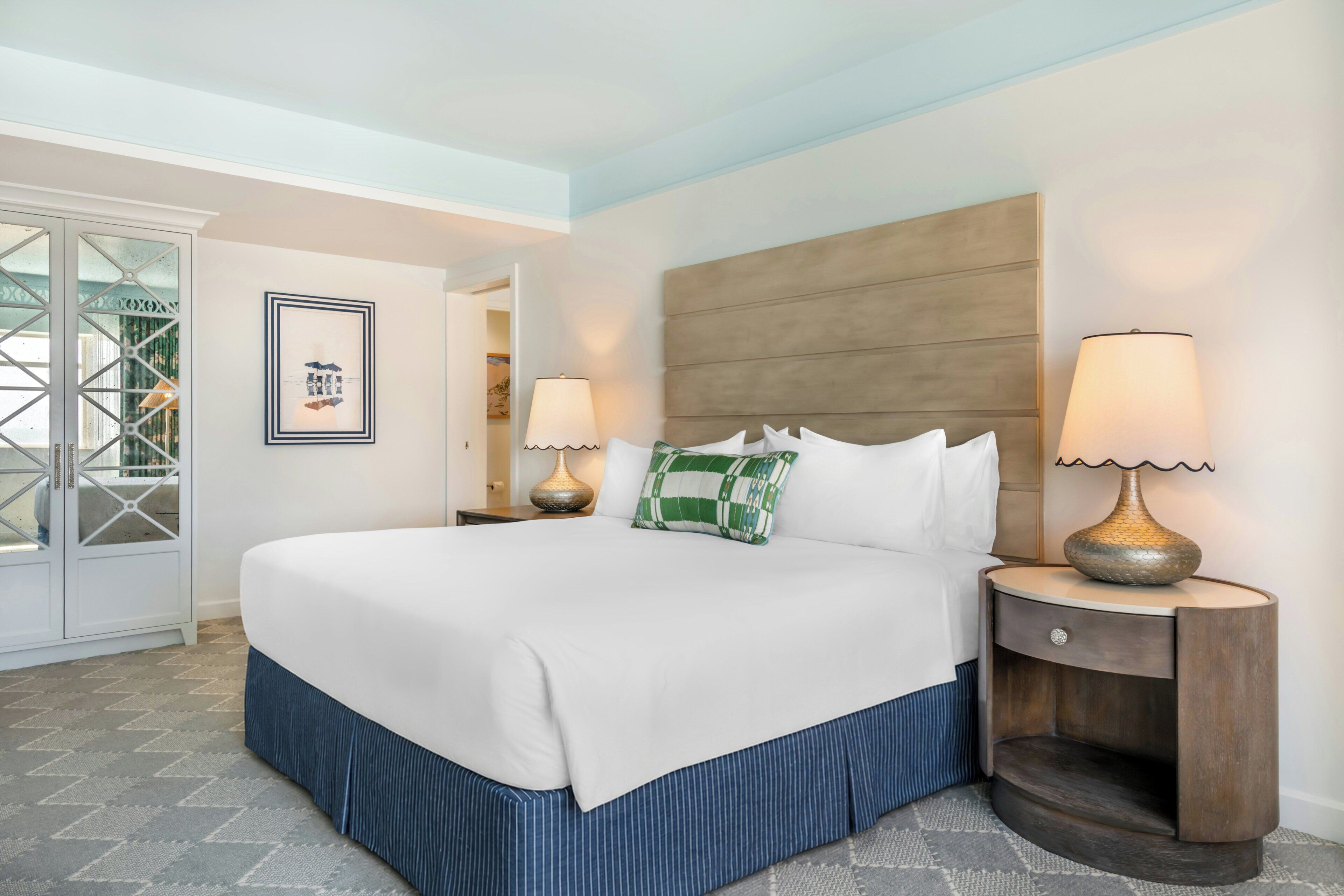 Habitación de hotel con una cama grande, una mesita de noche con lámparas, un cuadro en la pared y vistas al exterior a través de una puerta.