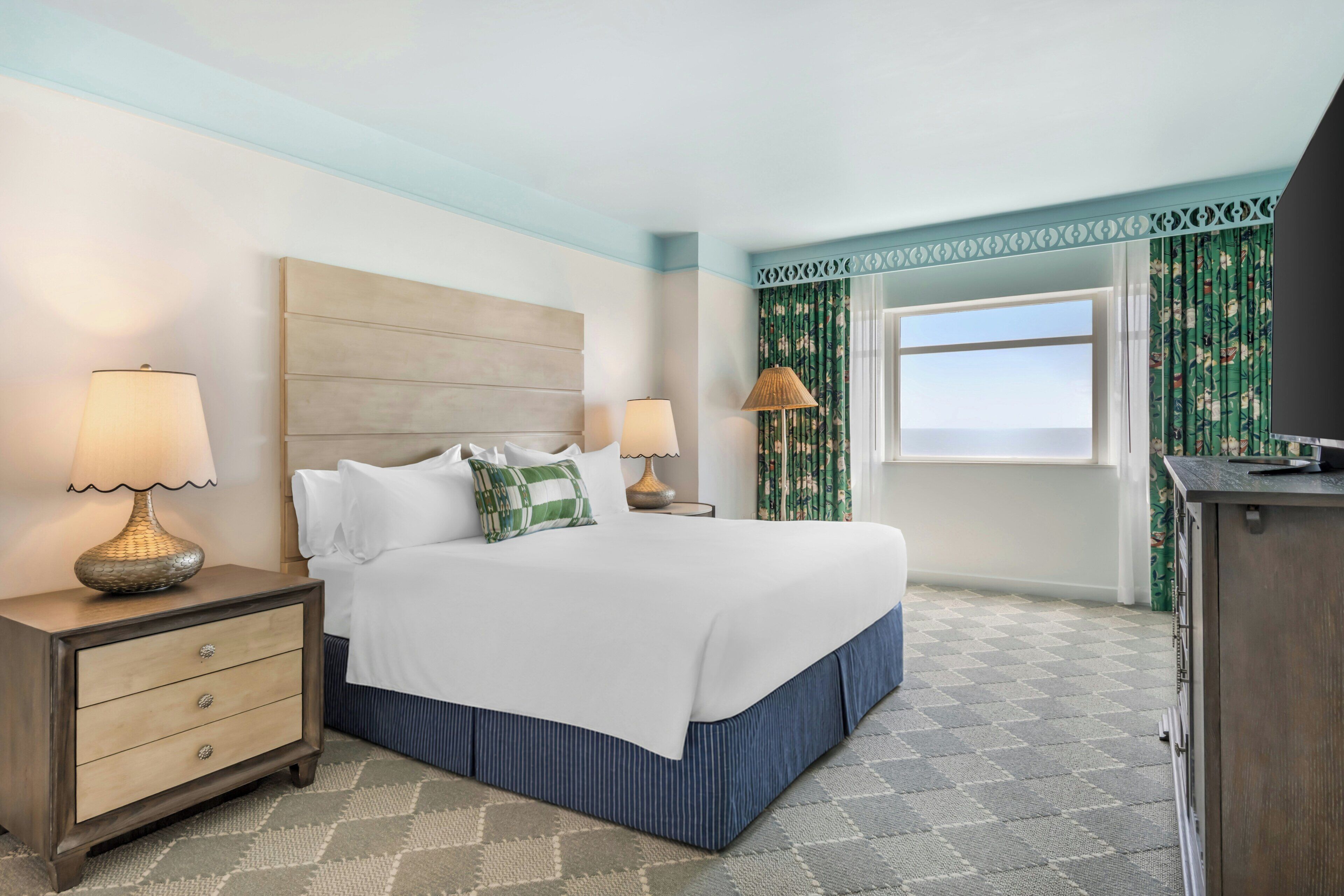 A hotel room with a bed, two bedside tables, a dresser, a window with curtains, and a view.