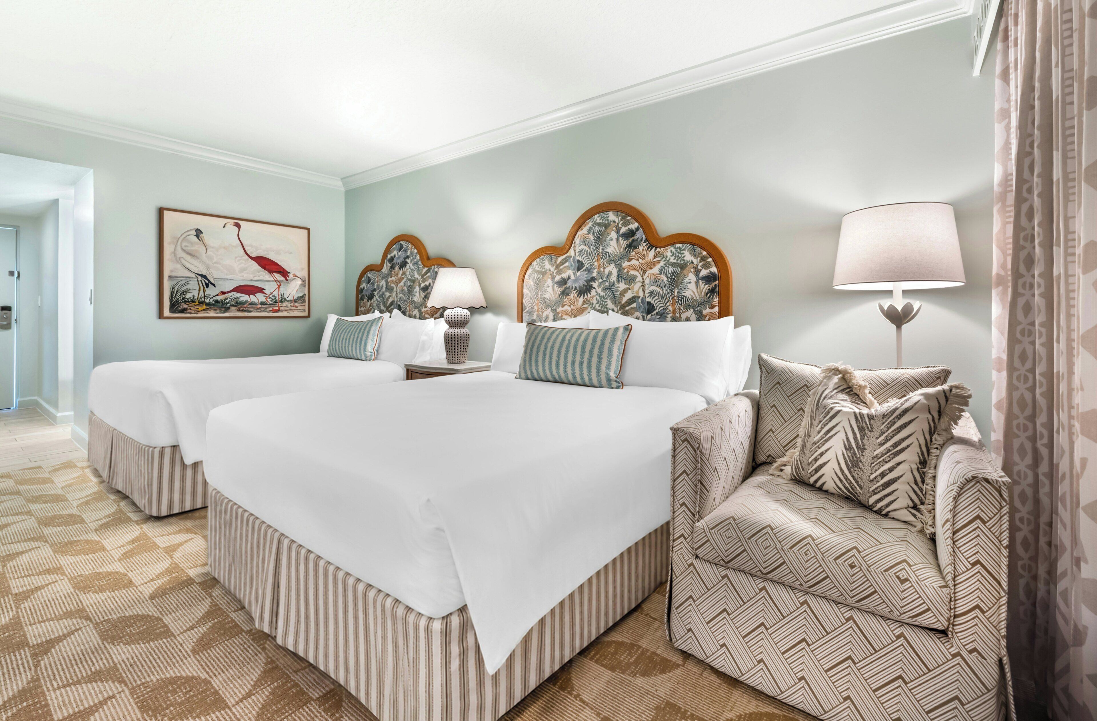 A hotel room with two beds, a patterned armchair, a painting, and a lamp.