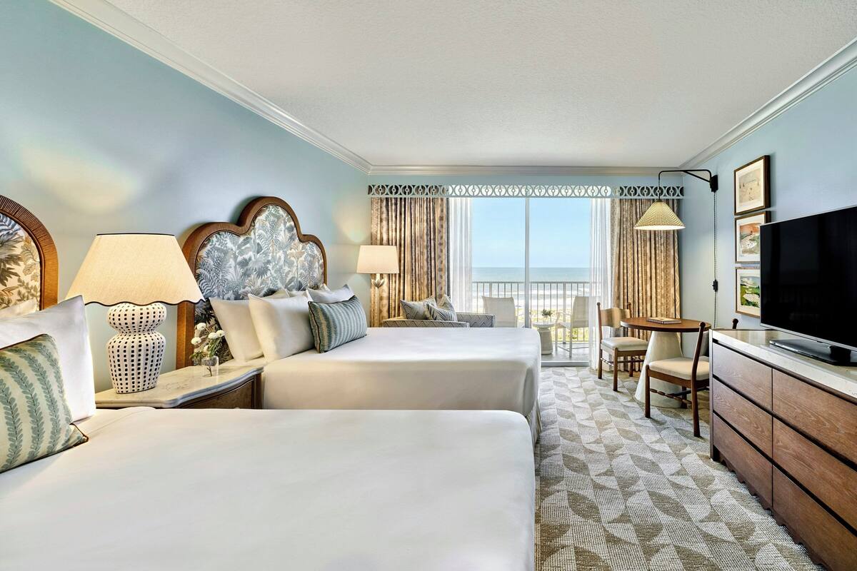 A hotel room with two beds, a TV, a dining table with chairs, and a balcony with curtains.