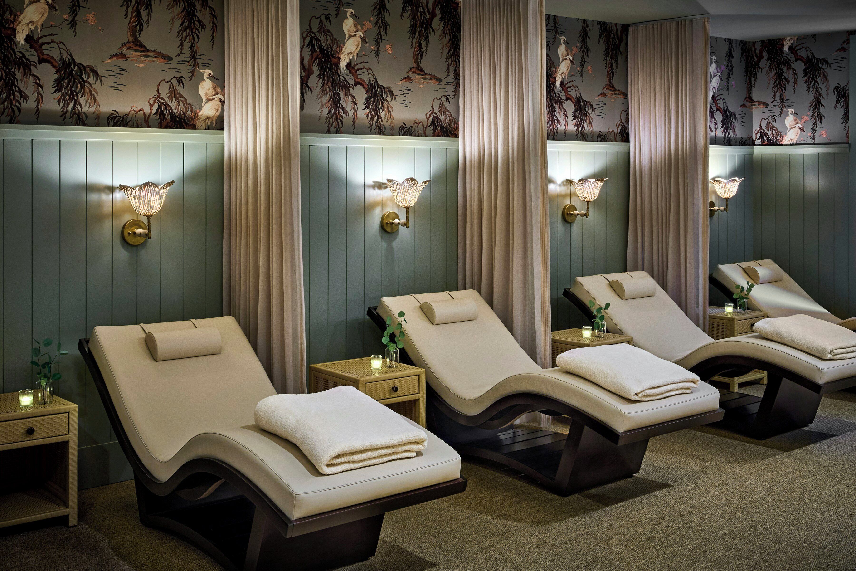 A spa room with four reclining chairs, each with a towel, a small table with a candle, and wall-mounted lamps.