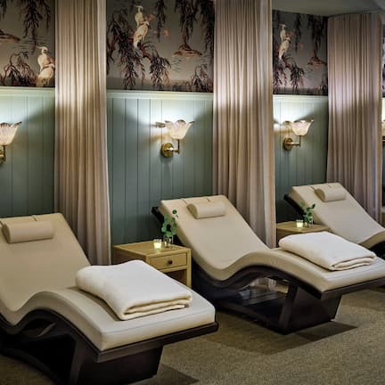 A spa room with four reclining chairs, each with a towel, a small table with a candle, and wall-mounted lamps.