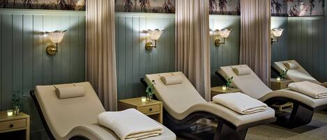 A spa room with four reclining chairs, each with a towel, a small table with a candle, and wall-mounted lamps.
