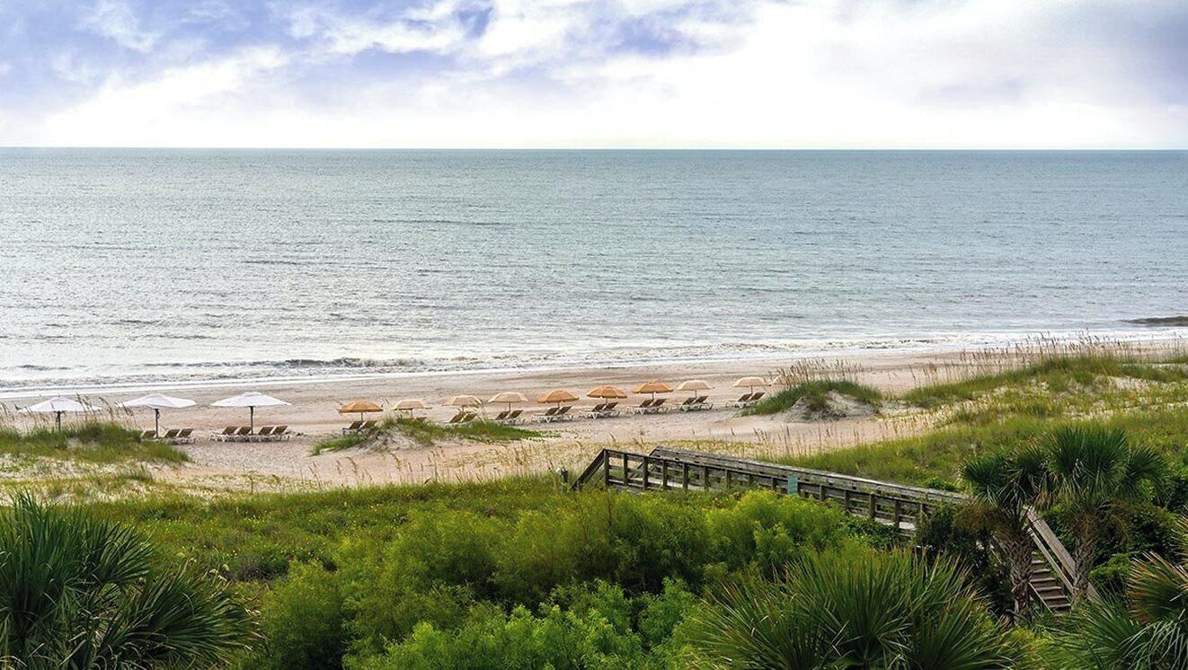 A beach with umbrellas and lounge chairs, a wooden boardwalk, and a view of the ocean.