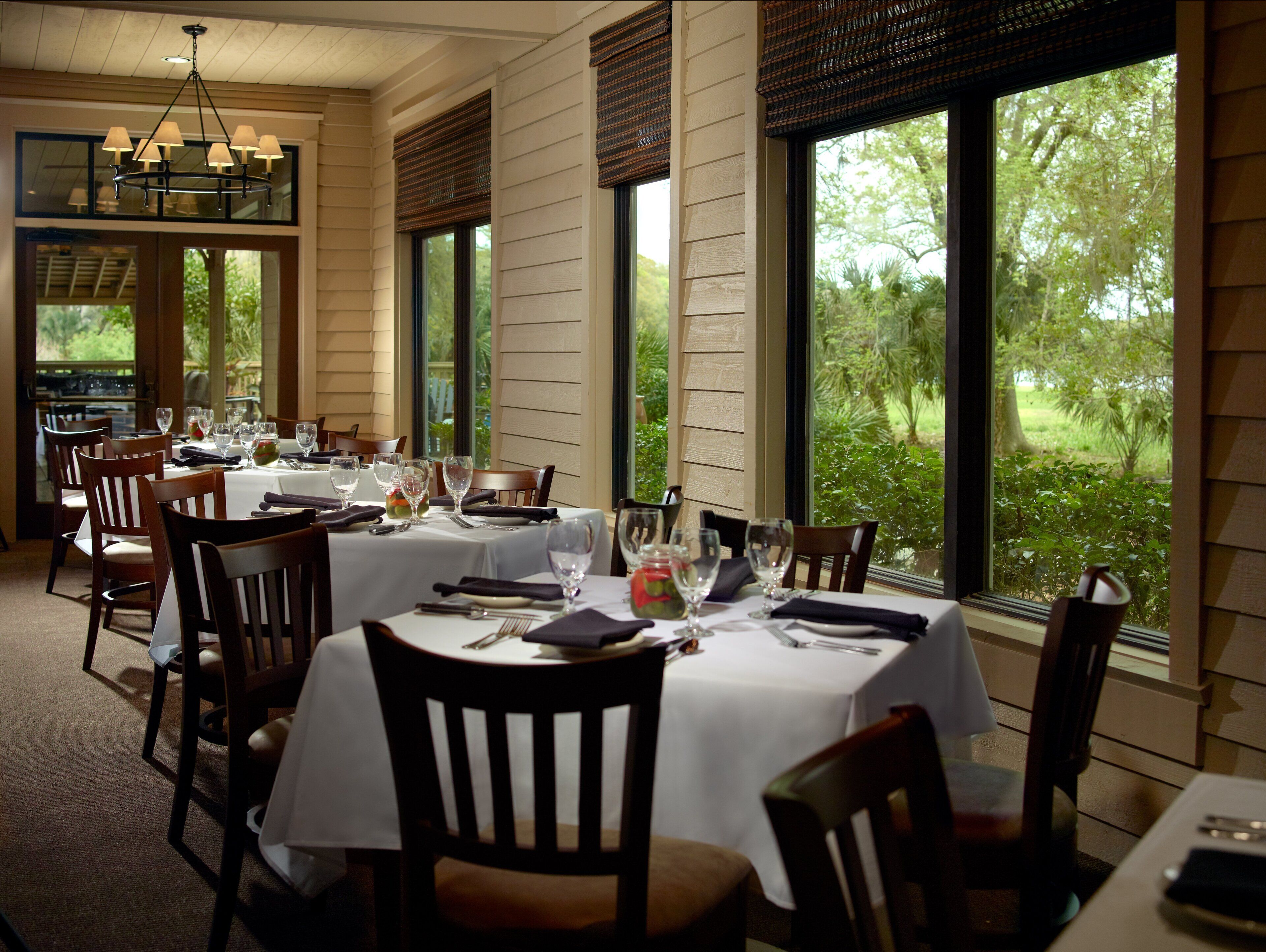 A dining area with tables set for a meal, large windows with views of greenery, and a chandelier.