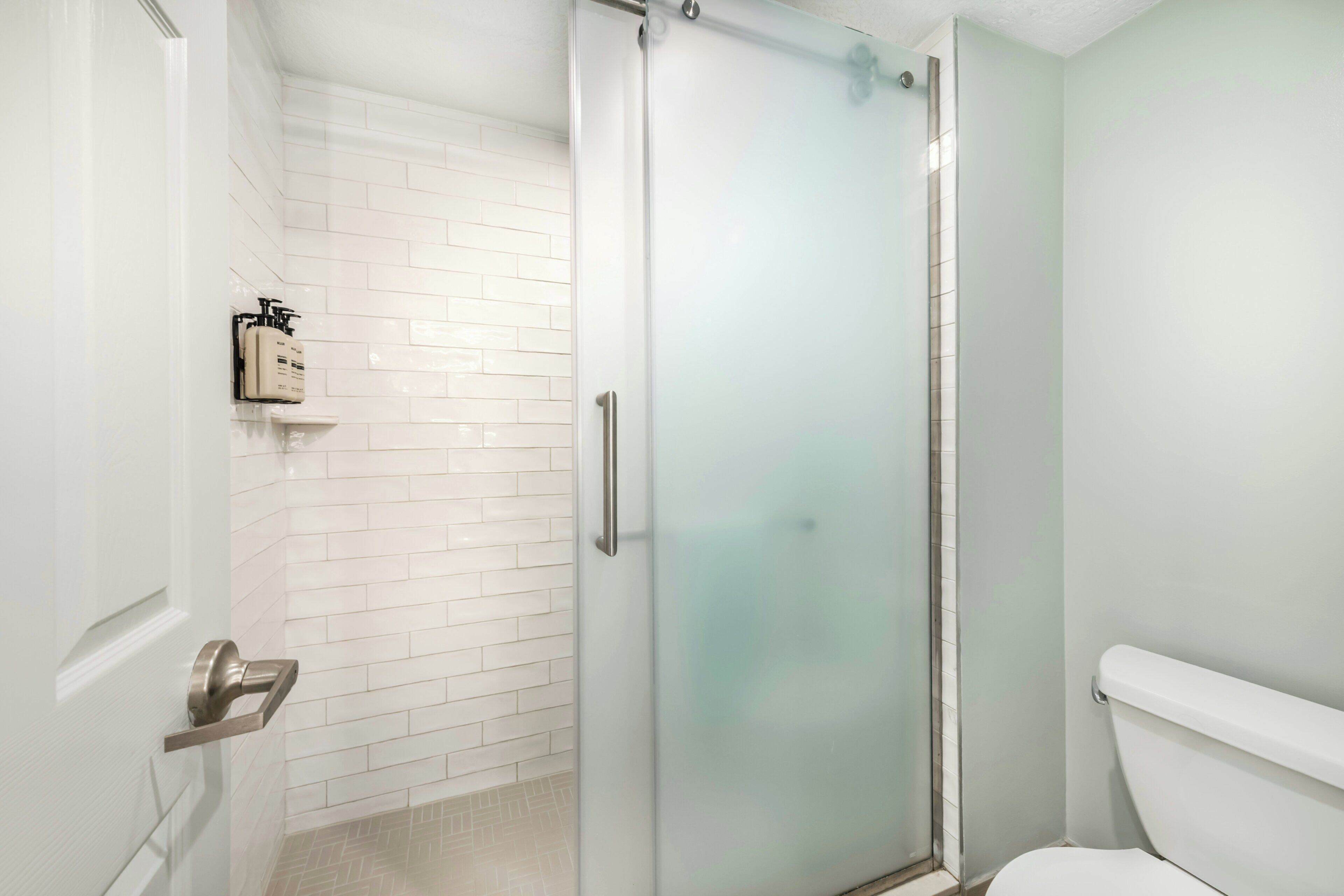 Un baño con mampara de vidrio, inodoro blanco y pared con azulejos blancos.