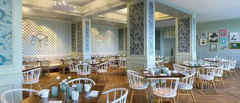 A spacious dining area with white chairs and tables, a patterned wall, and a ceiling with a central light fixture.