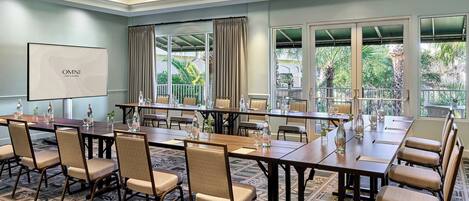 Un salón de conferencias con una larga mesa, sillas, una pantalla de proyector y ventanas grandes con cortinas.