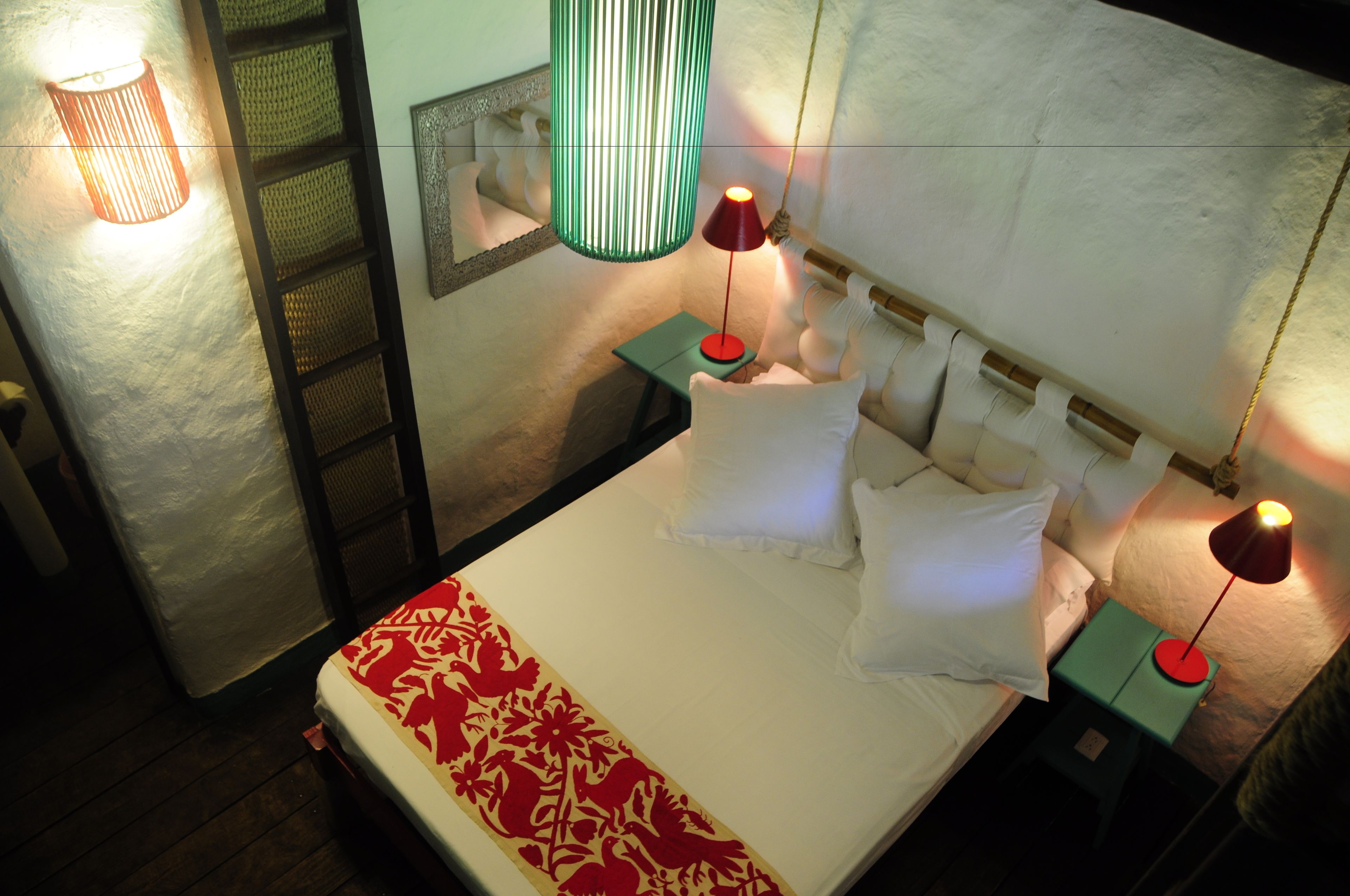 Una cama bien hecha con sábanas blancas y un corredor rojo estampado, dos mesitas de noche con lámparas, una lámpara colgante verde y una pared con textura.