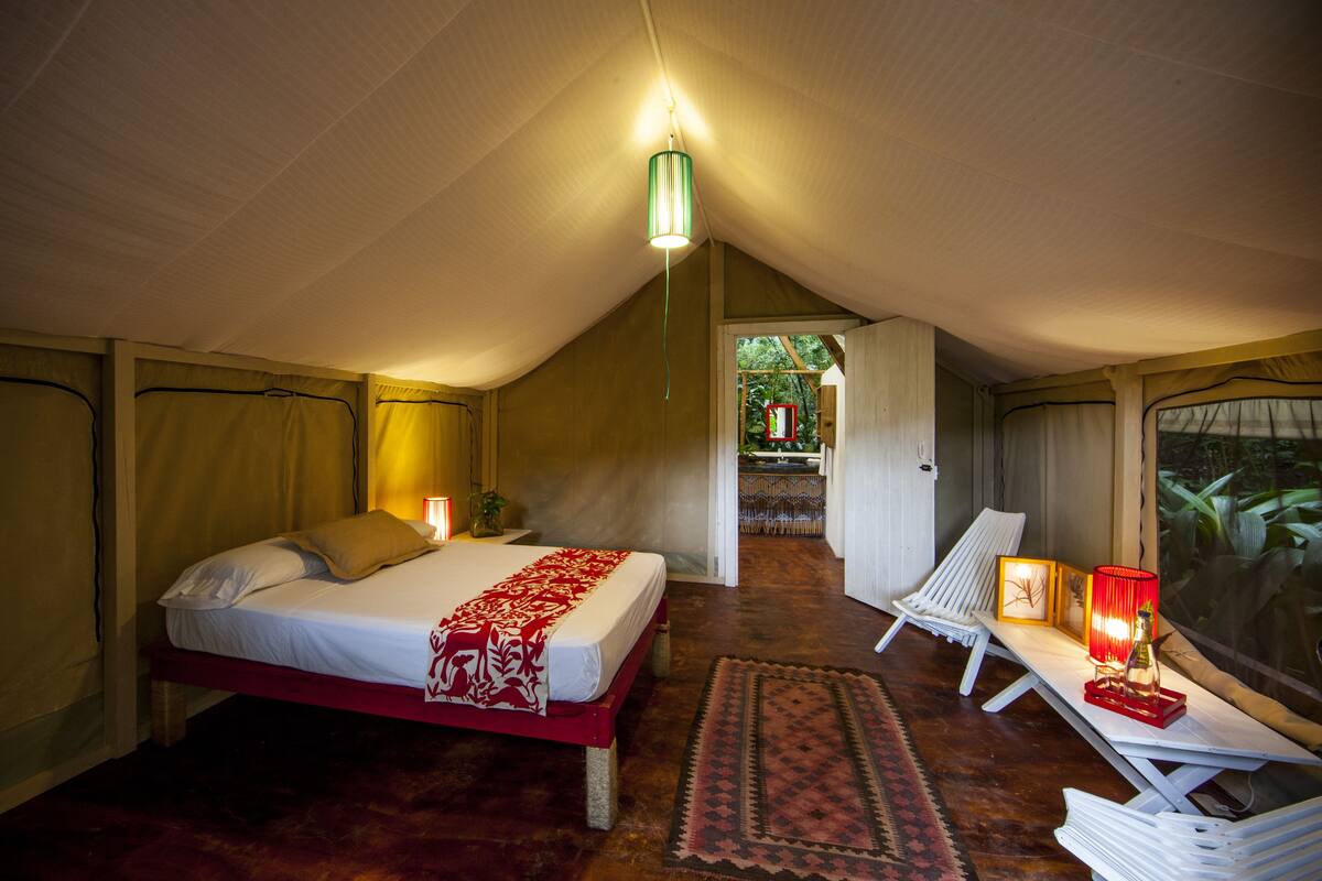 A cozy tent-style room with a bed, a lounge chair, and a small table with a lamp.