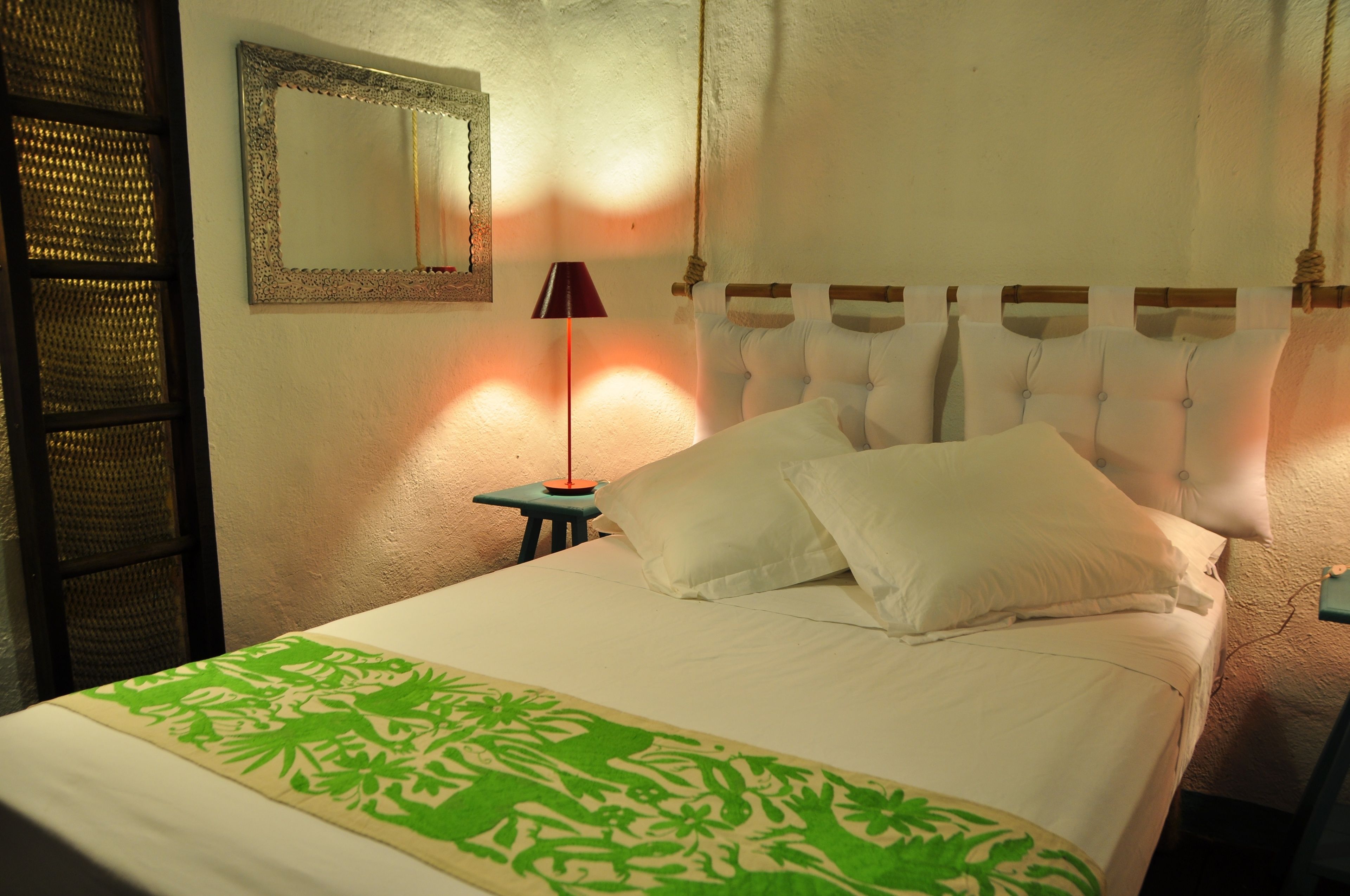 Una cama bien hecha con sábanas blancas y un plaid verde estampado, una lámpara de noche y un espejo en la pared.
