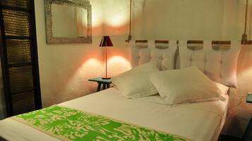 Una cama bien hecha con sábanas blancas y un plaid verde estampado, una lámpara de noche y un espejo en la pared.