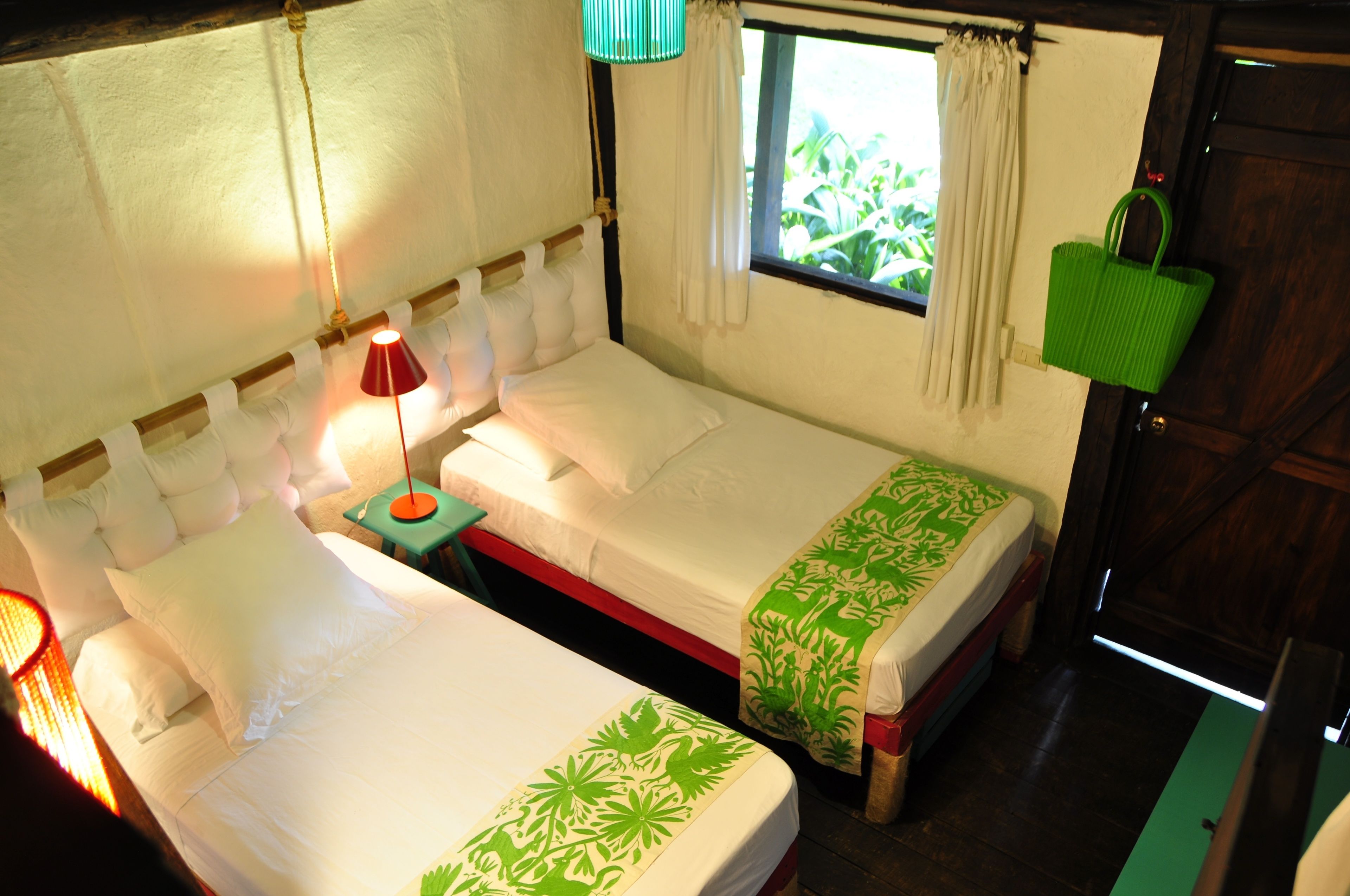 A room with two beds, a window with curtains, a lamp, and a green bag hanging on the wall.