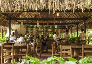 A rustic outdoor dining area with wooden tables and chairs, surrounded by lush greenery and a thatched roof.