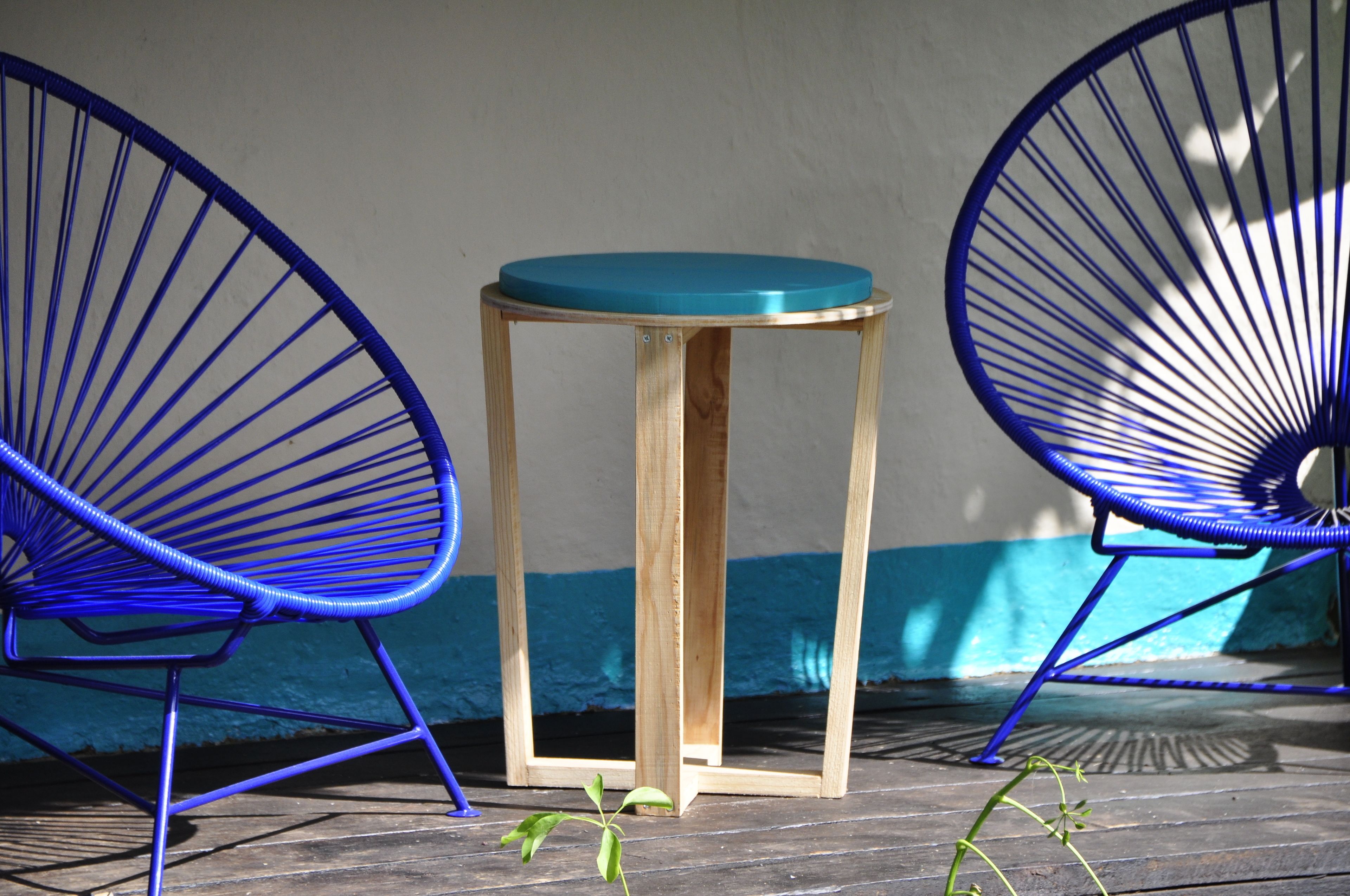 Dos sillas azules y una mesa de madera pequeña con tapa color verde azulado, colocadas frente a una pared con rayas azules y blancas.