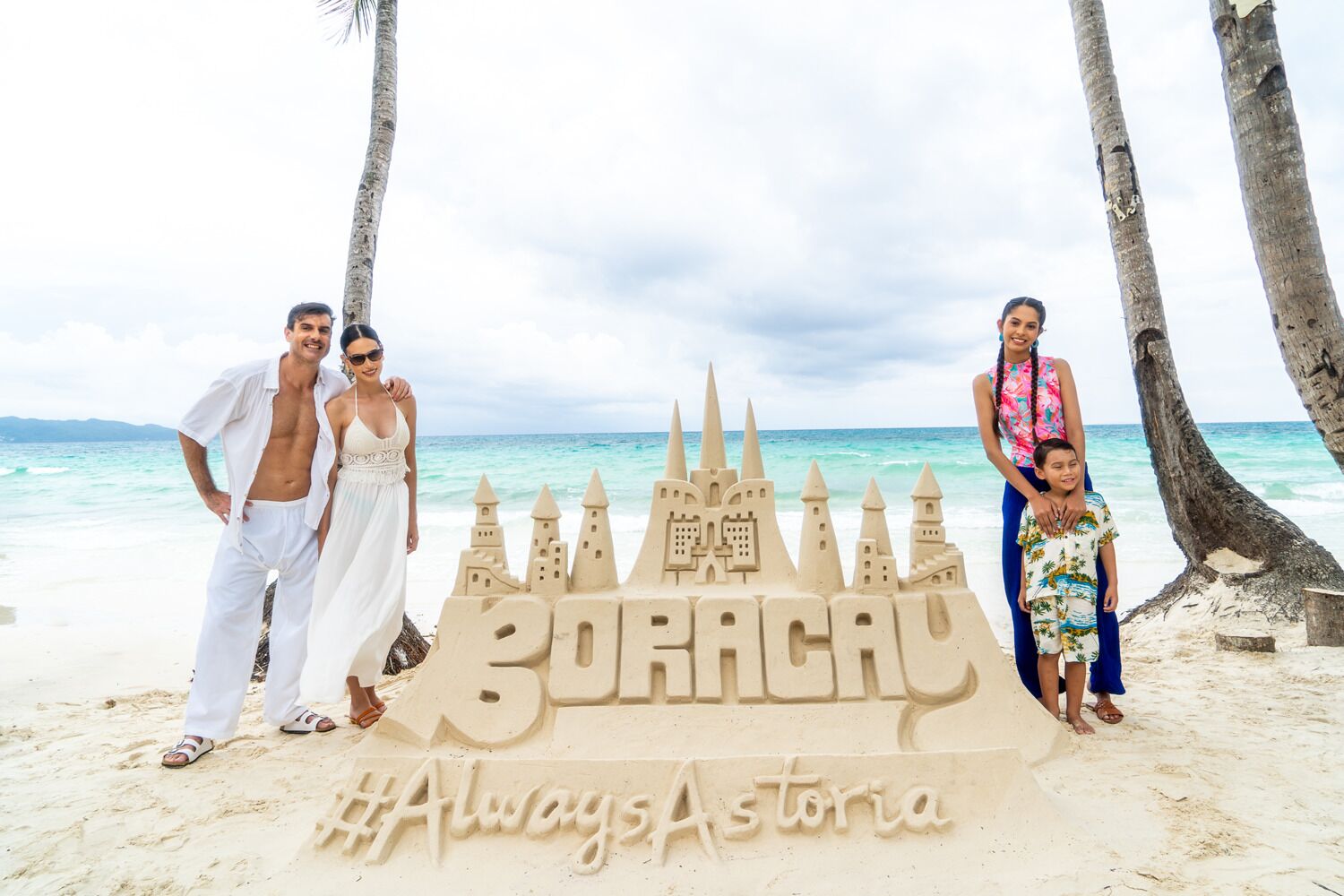 A family posing in front of a sandcastle with the text "BORACAY #AlwaysAstoria" on a beach.