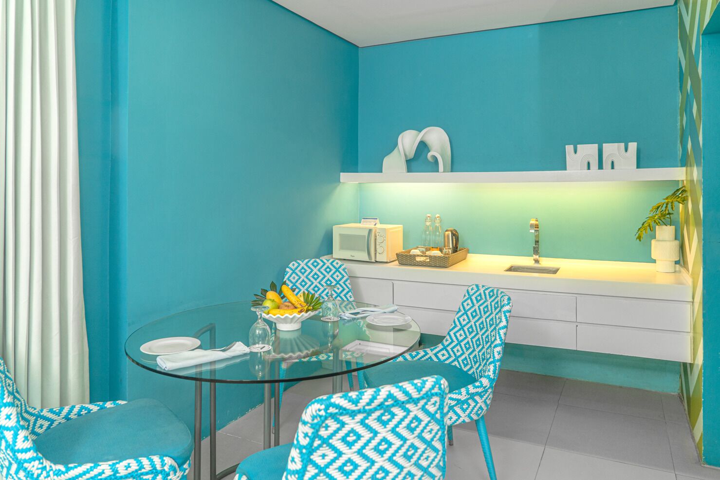 Una cocina moderna con una mesa de comedor de cristal, sillas azules con estampado y una encimera blanca con microondas y tostadora.