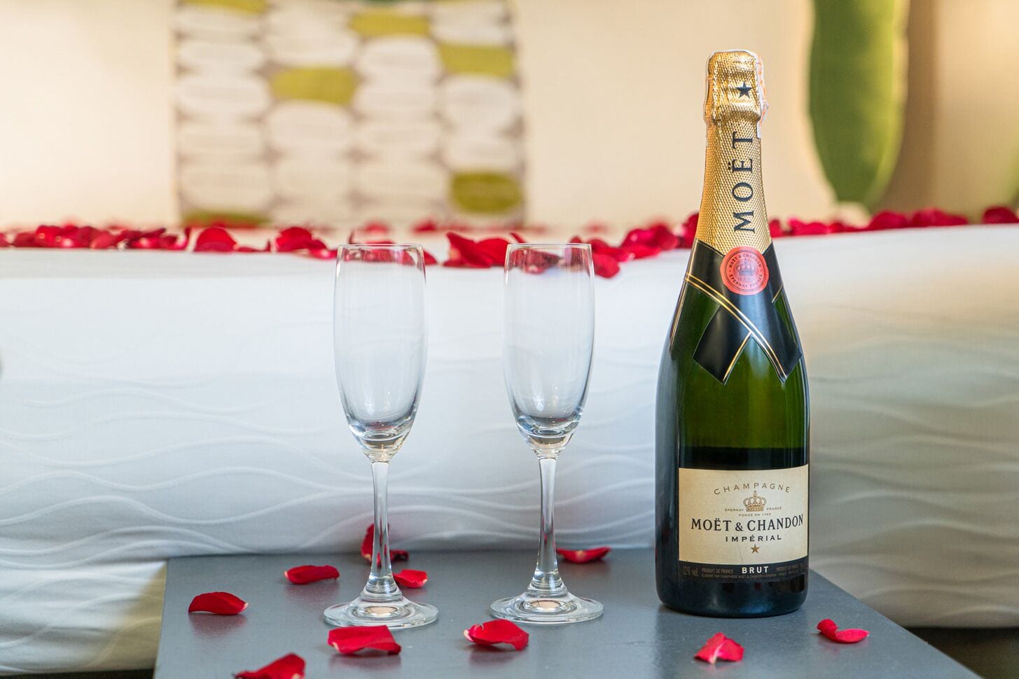 Eine Flasche Moët & Chandon Champagner und zwei Gläser auf einem Tisch mit Rosenblättern, vor einem Bett mit einem weiß-grünen Kopfende.