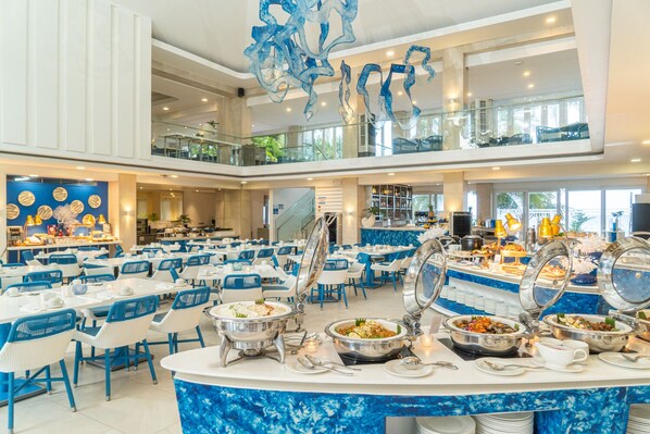 Amplia zona de comedor de hotel con varias estaciones de buffet, sillas blancas y azules, y un mostrador central de servicio.
