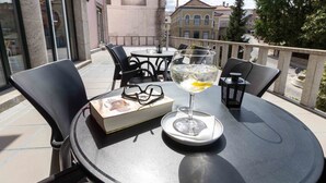 Een ronde tafel met een glas witte wijn, een boek en een paar brillen op een balkon met uitzicht op een straat met gebouwen en bomen.