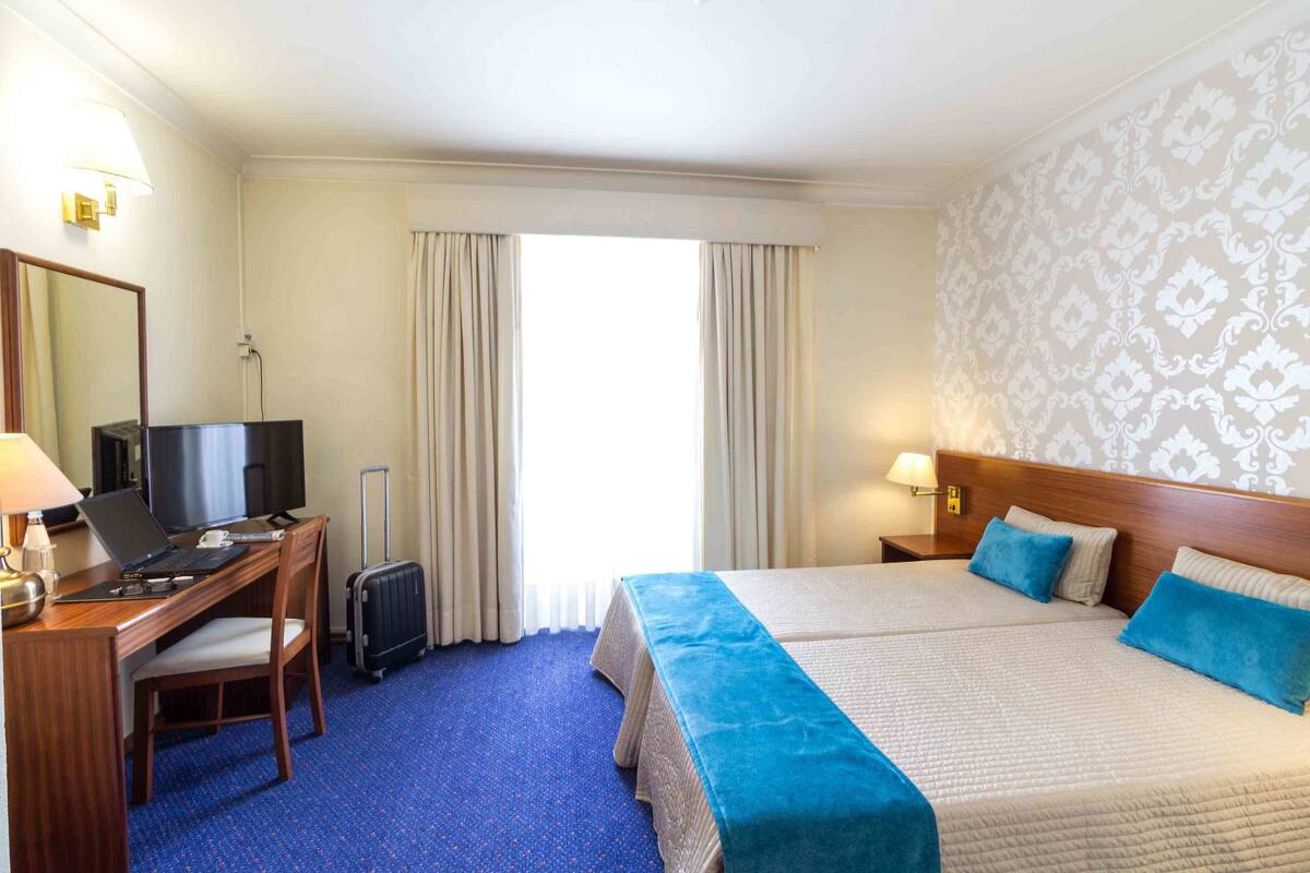 A hotel room with a bed, a desk with a laptop, a television, and a suitcase.