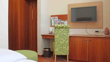 A hotel room with a bed, a desk with a flat-screen TV, a trash can, and a cabinet.
