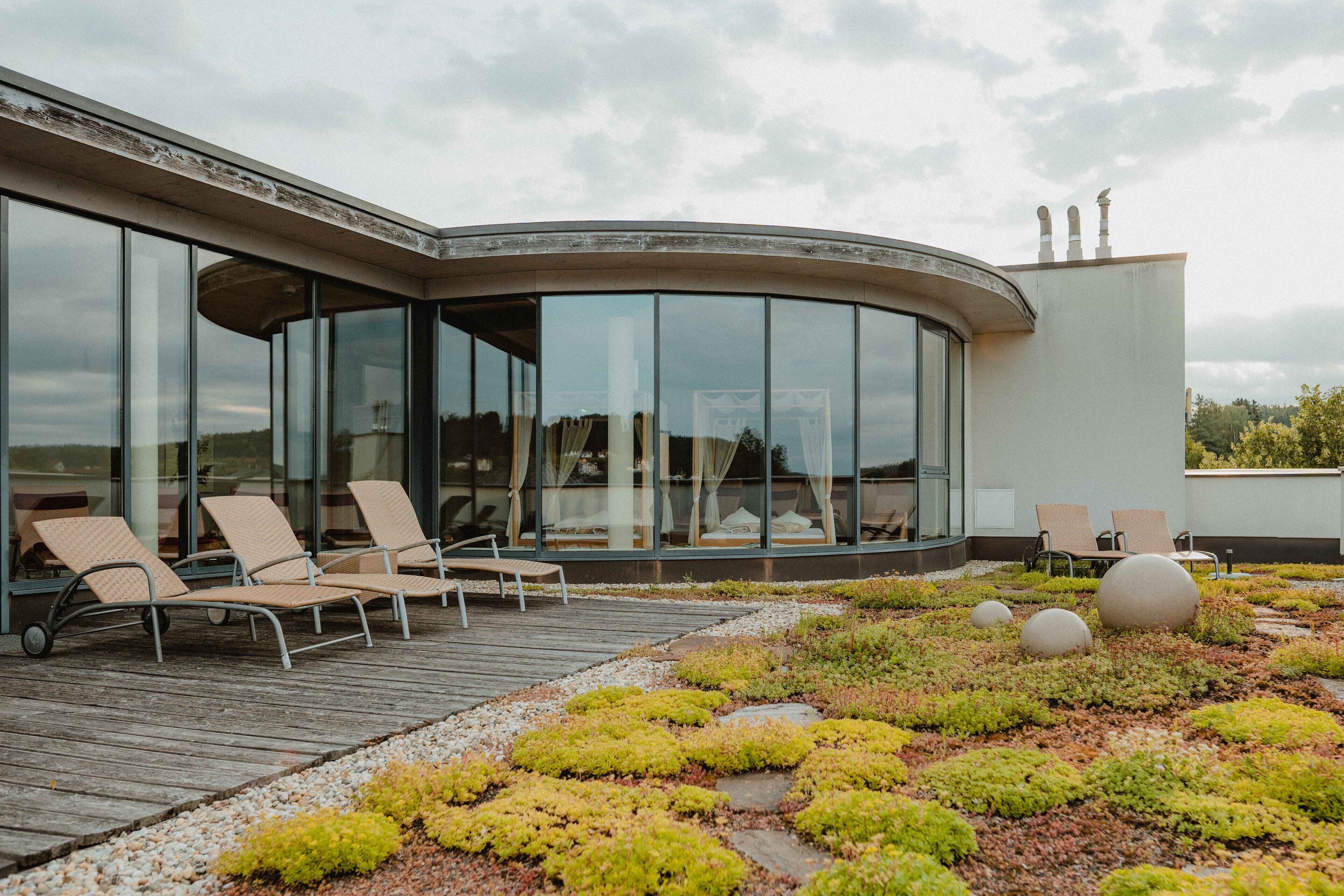 Un edificio moderno con amplios ventanales, zona de estar exterior con sillones y un jardín paisajístico con musgo y piedras.